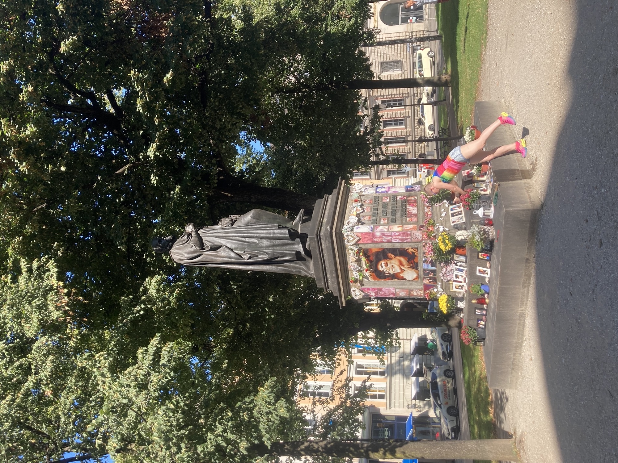 Statue auf einem hohen Sockel, der Sockel ist mit vielen Fotos von Michael Jackson, Herzen und Blumen dekoriert, die Statue ist nicht Michael Jackson, rechts am Sockel richtet eine bunt gekleidete Person die Fotos gerade