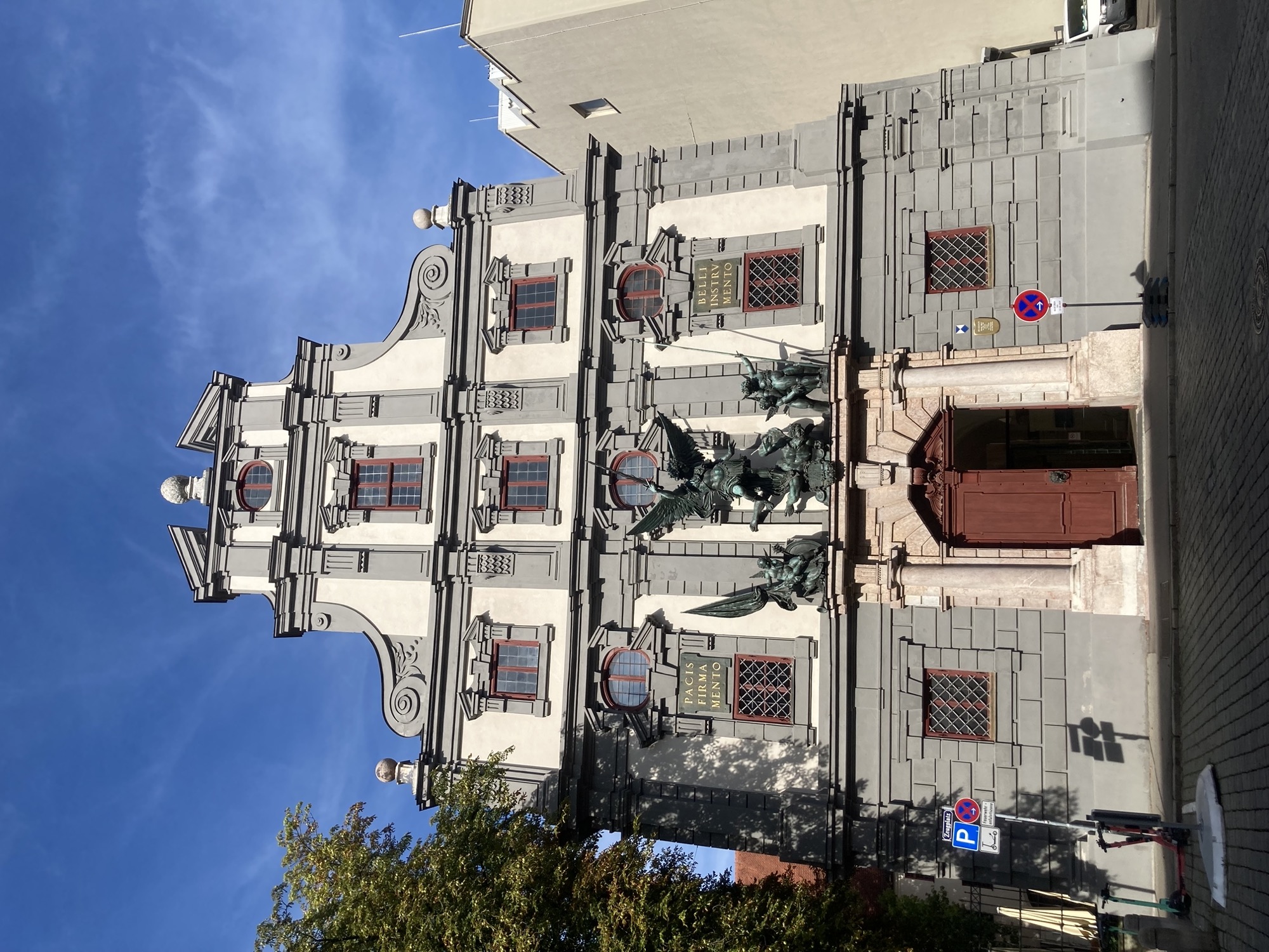 Augsburger Zeughaus, Fassade im Manierismus-Stil, verschiedene Fensterformen, graue Verzierungen um die Fenster herum, über dem rötlich-braunen Tor wacht eine Statuengruppe, links und rechts davon Marmortafeln mit lateinischer Aufschrift, links: PACIS FIRMA MENTO, rechts, BELLI INSTRV MENTO