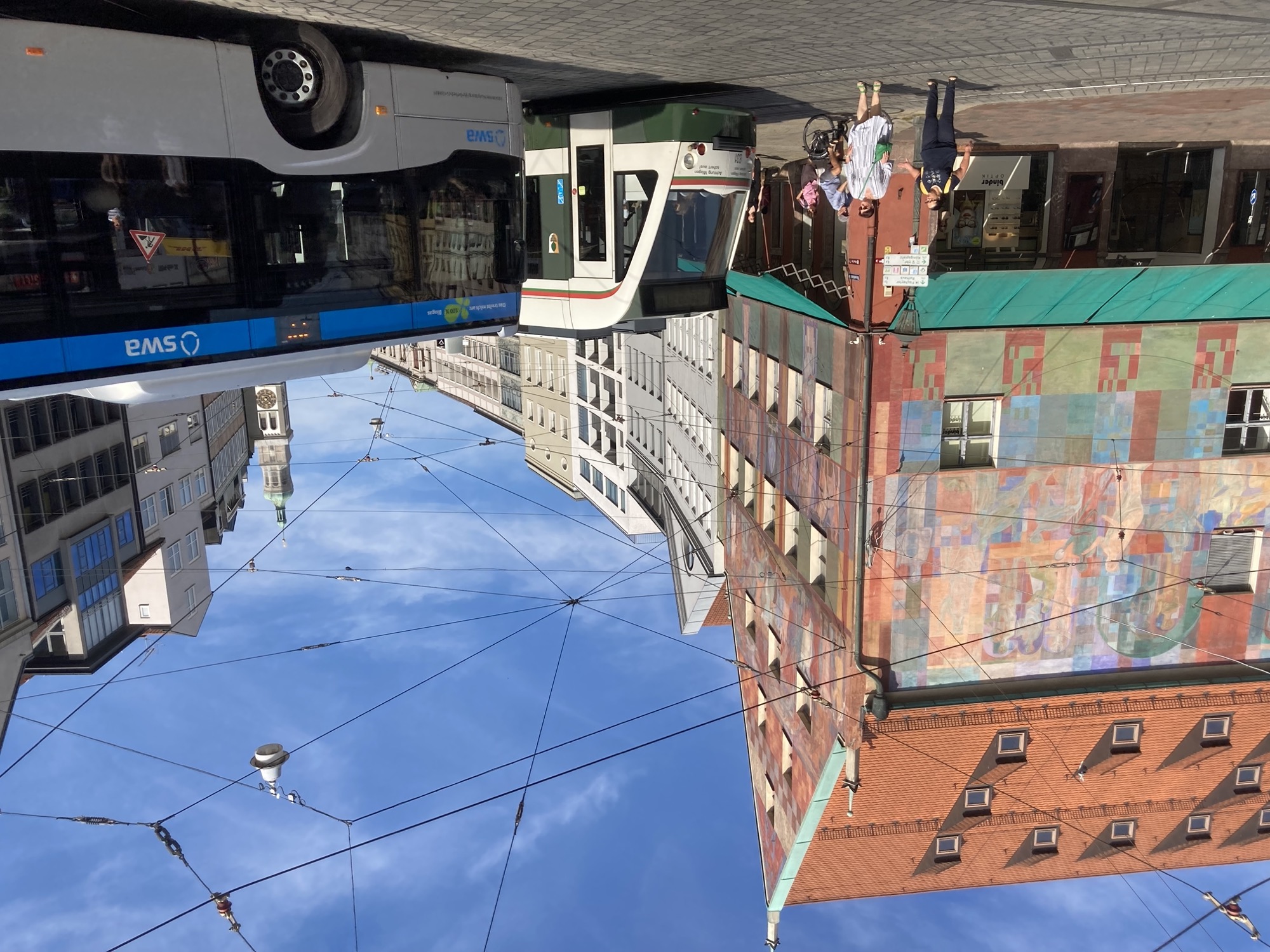 Verkehr vor dem Weberhaus, ein Haus mit Spitzdach und zahlreichen Dachgauben, bunt bemalte Fassade, von rechts nähern sich eine Straßenbahn und ein Bus, vor der Straßenbahn blockieren zwei Personen das Gleis