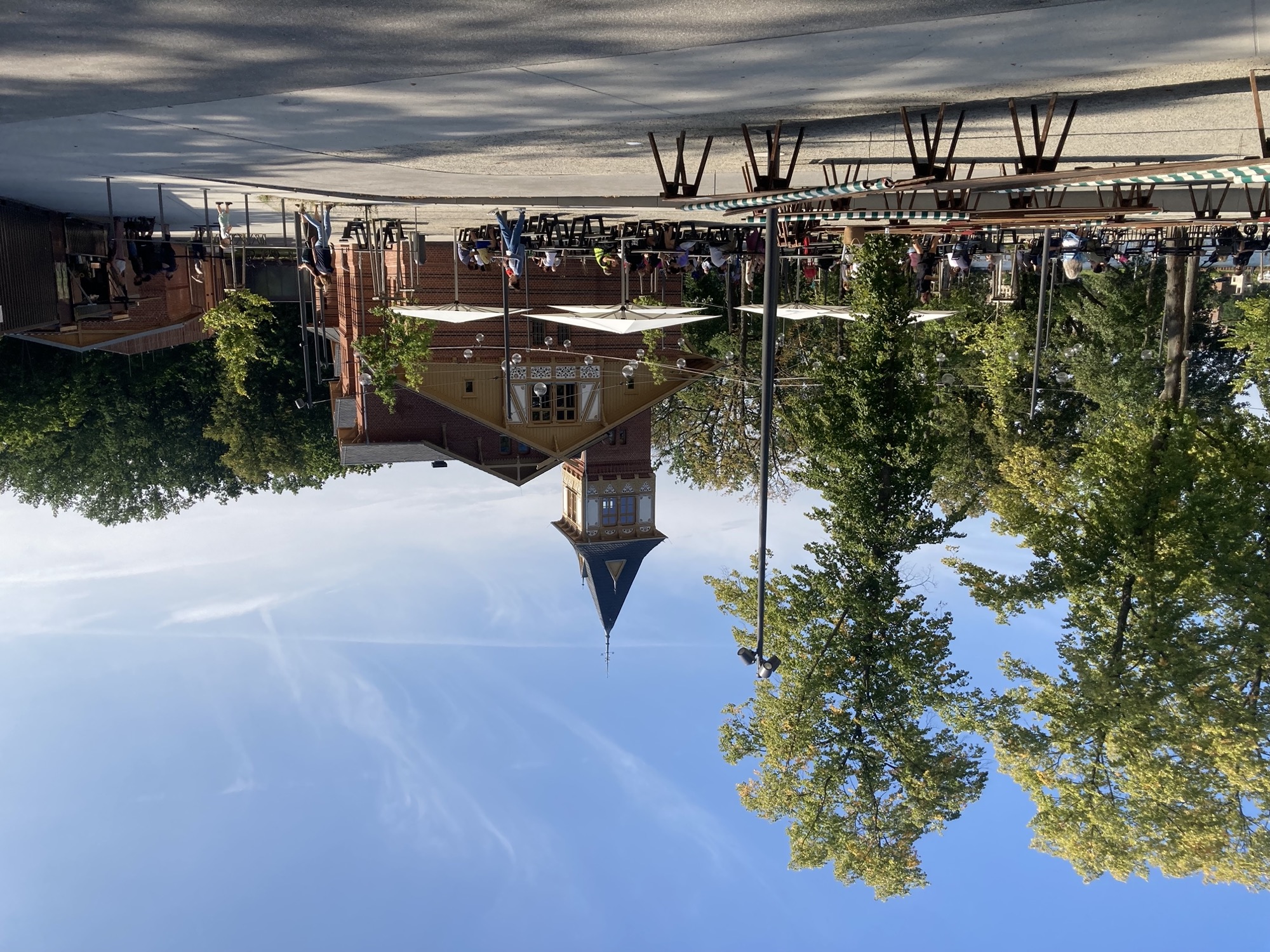 Biergarten neben dem Eierhäuschen, vor blauem Himmel mischt sich Sonne mit Schatten, das Eierhäuschen ist ein schmuckes Geböäude mit einem Turm, rechts im Bild die Jausenstation mit einer kleinen Menschenschlange davor