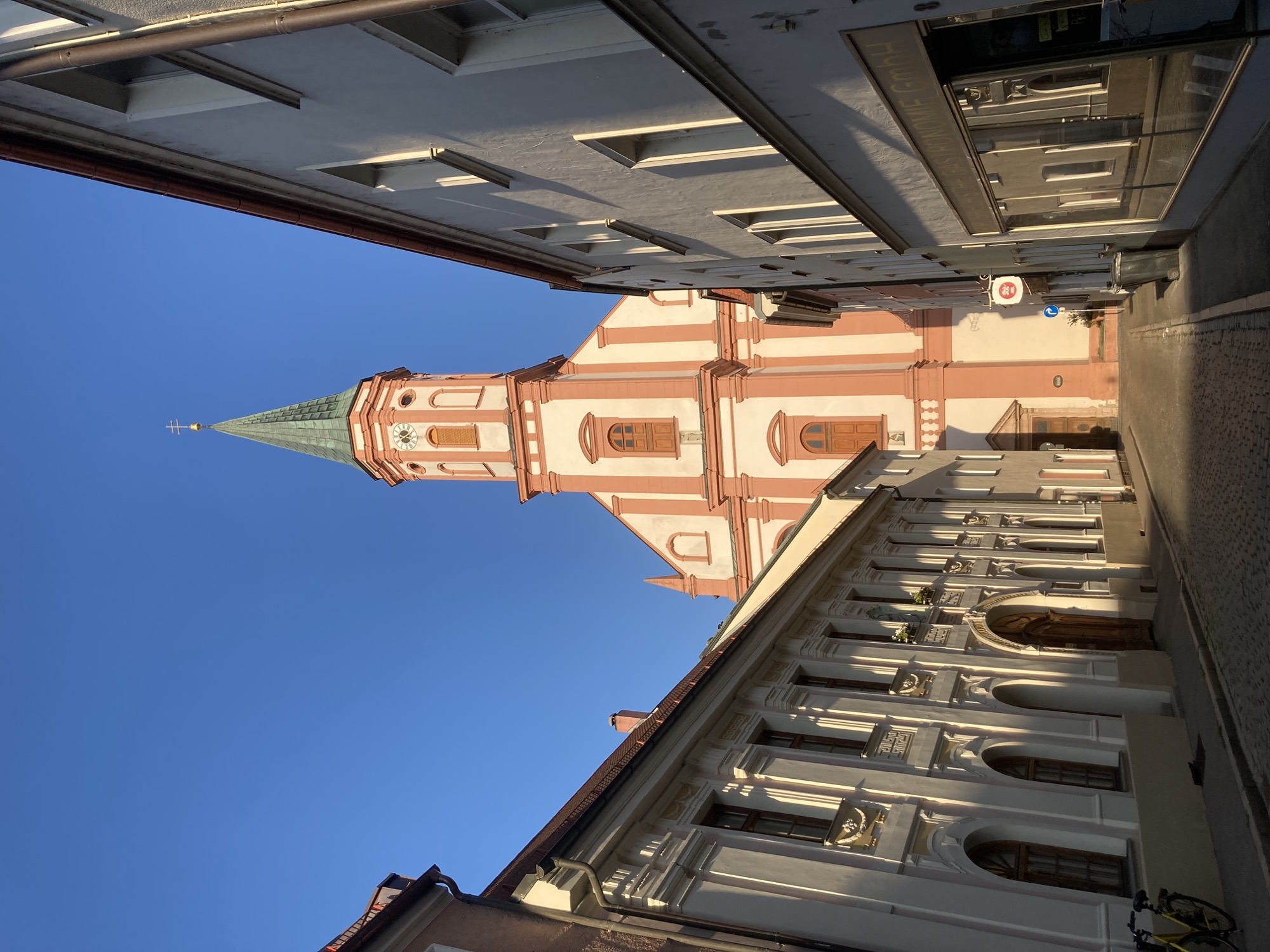 am Ende einer schmalen Gasse ragt ein Kirchturm in die Höhe, die Kirche ist hell mit rötlichen Säulen und Verzierungen, der Turm oben achteckig