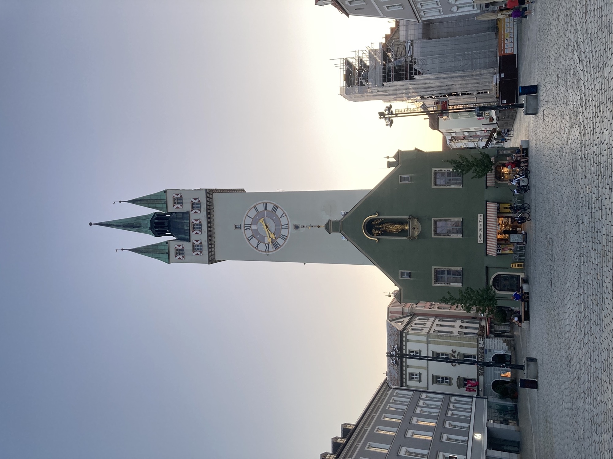 Stadtturm Straubing, von hinten im Abendlicht beleuchtet, im Vordergrund das Gebäude um den Stadtturm, grün gestrichen, zwei Stockwerke, im Erdgeschoss heute ein Eisgeschäft, im Giebel eine große goldene Heiligenstatue