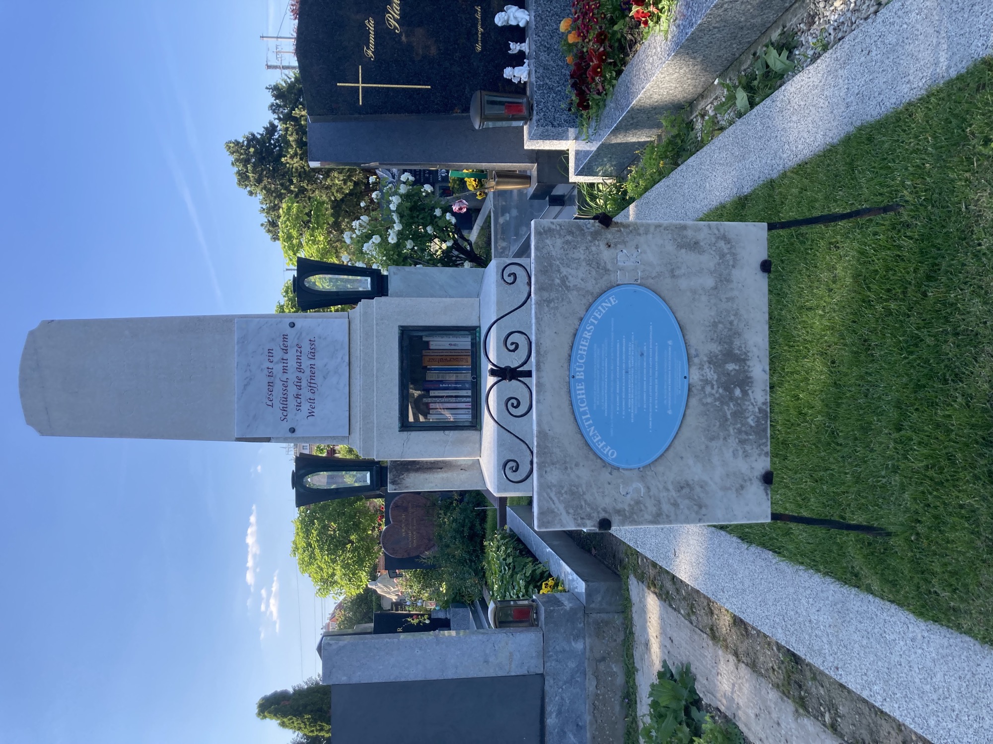 Grabstein auf dem Friedhof, auf dem Grabstein ist eine Marmortafel mit diesem Text befestigt: „Lesen ist ein Schlüssel, mit dem sich die ganze Welt öffnen lässt.“, darunter ist im Grabstein ein Fach eingelassen, in dem eine Reihe Bücher steht, auf der Grabfläche davor ein blaues rundes Schild mit der Beschriftung „Öffentliche Büchersteine“ und darunter weiterem Text, der aber nicht lesbar ist