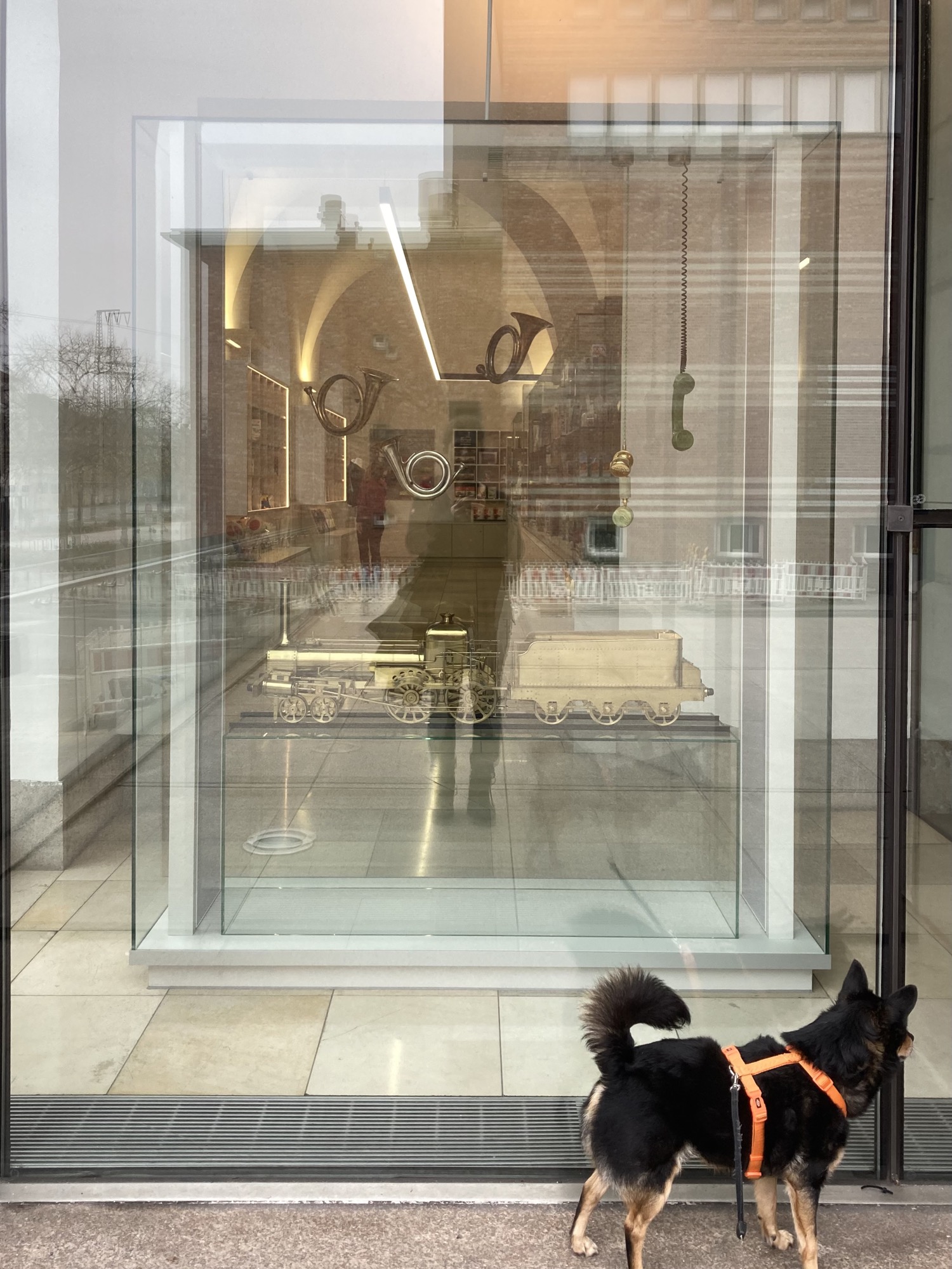 Detailaufnahme eines Schaufensters, drinnen in einem Glaskasten ein goldener Zug, von oben hängen Posthörner und Telefonhörer herab, rechts unten im Bild ein schwarzer Hund