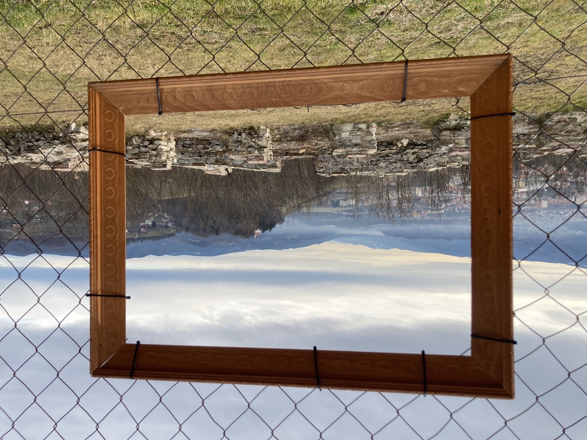 Blick durch einen Bilderrahmen, der ein Loch in einem Zaun einrahmt, innerhalb des Bilderrahmens im Vordergrund Überreste von Wänden eines Schlosses, weiter hinten ein Hügel mit einer Kirche darauf, im Hintergrund nebelverhangene Berge