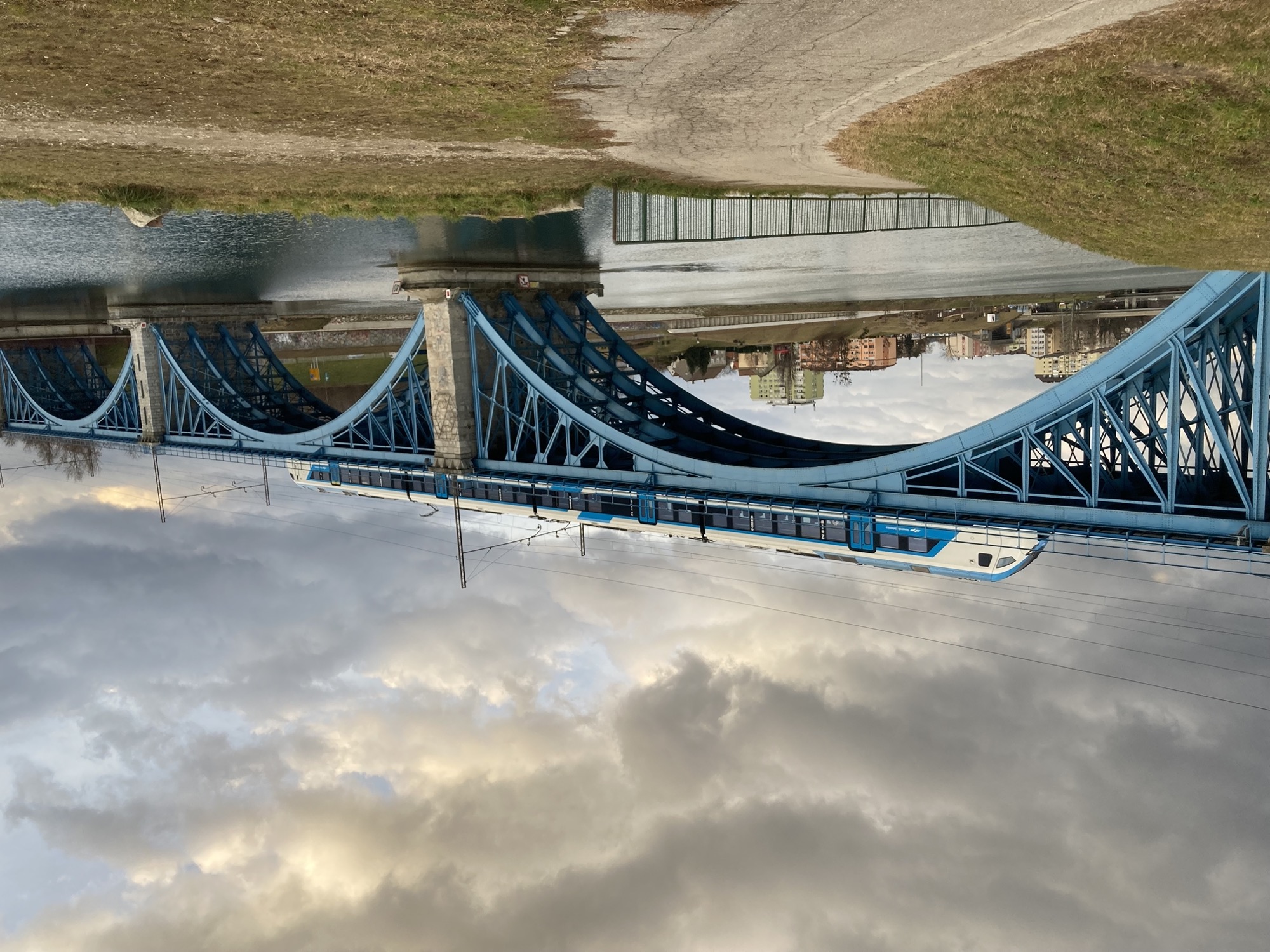 über einen Fluss spannt sich eine blaue Eisenbahnbrücke, auf der Brücke fährt ein blau-weißer Zug gerade schräg von rechts unten im Bild nach links oben