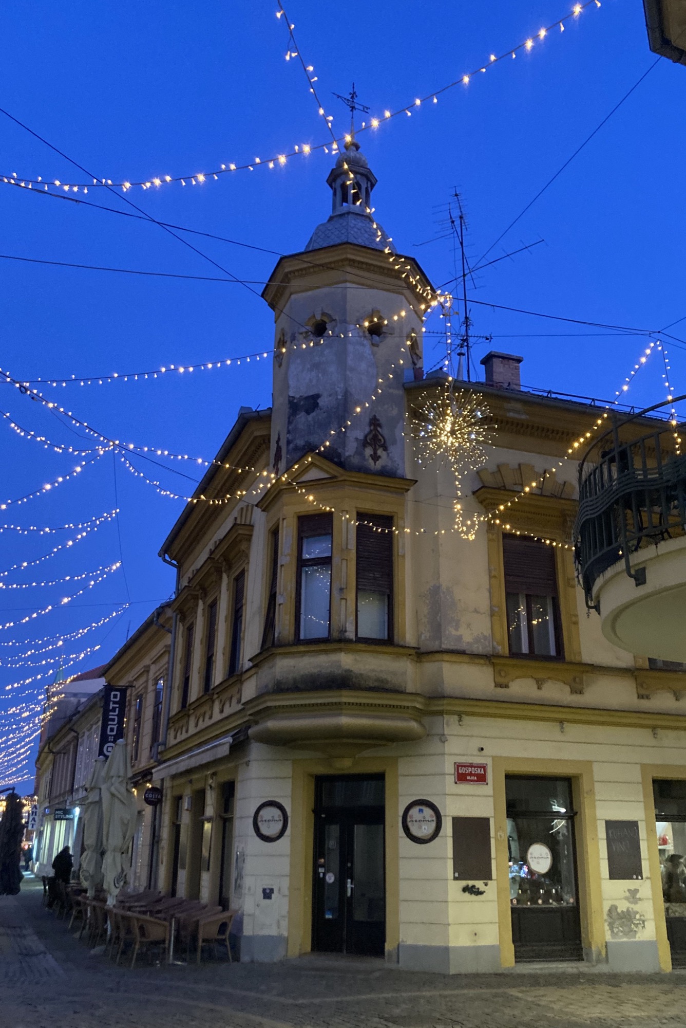 ein Eckhaus in einer Fußgängerzone am Abend vor dämmrigem blauem Himmel, das Haus hat einen Erker und einen Turm mit einem Wetteranzeiger oben drauf, vor dem Haus und in der Gasse links am Bildrand hängen Lichterketten