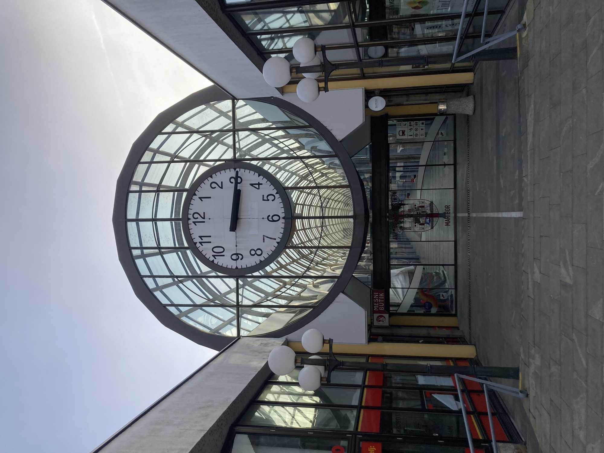 zentraler Busbahnhof in Maribor, zwischen zwei Längsgebäuden wölbt sich eine halbkreisförmige Dachkonstruktion, über dem Eingang mit Schiebetüren hängt zentral eine große Uhr, sie zeigt 15.15 Uhr an