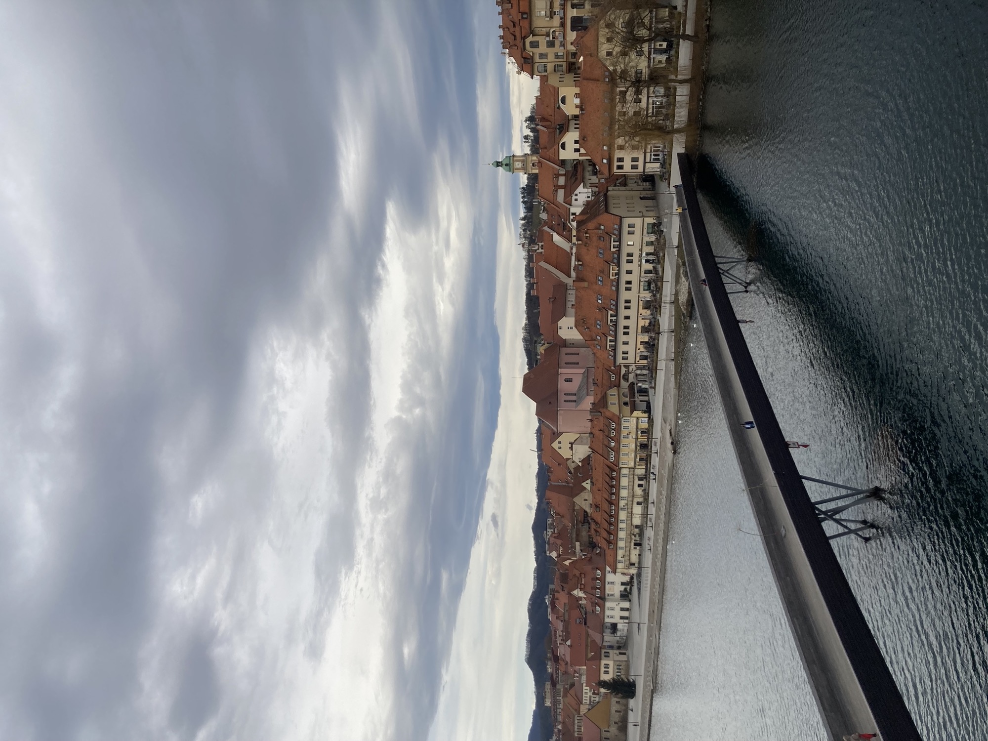 Ausblick über die Drau auf die Altstadt von Maribor, im Vordergrund eine Fußgänger:innenbrücke, dahinter die gelben Häuser am Ufer der Drau, weiter hinten überragt ein Kirchturm die Altstadt