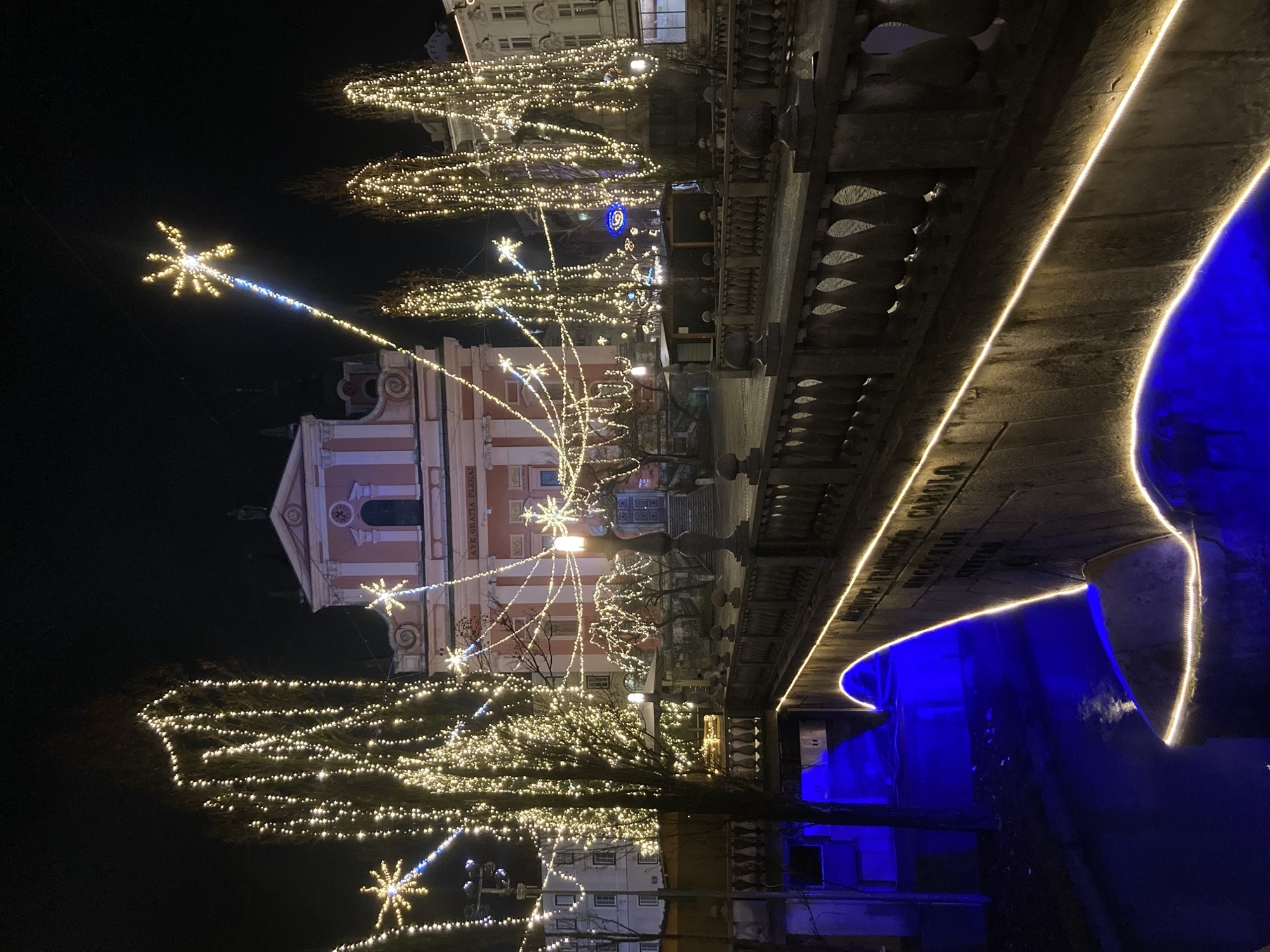 Nachtaufnahme, eine Brücke spannt sich quer durch das Bild von rechts unten nach links oben, der Fluss unter der Brücke ist blau beleuchtet, am Ende der Brücke ein Platz, der mit vielen Lichterketten überspannt ist, dahinter das Portal einer Kirche