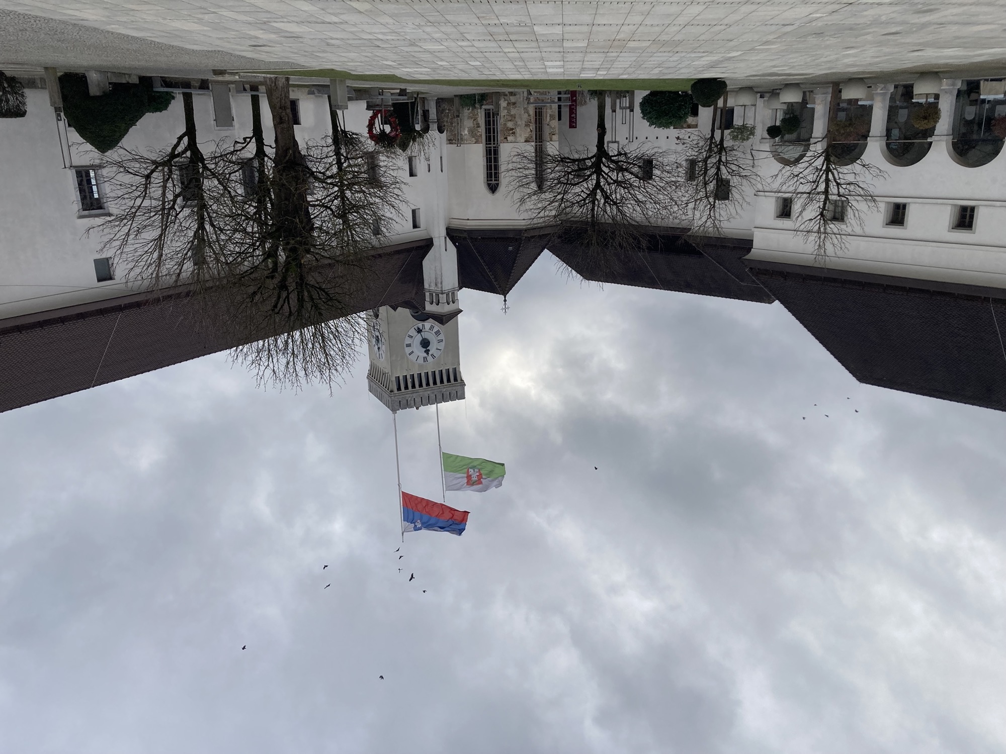 Innenhof im Schloss, über dem Uhrturm wehen die Flaggen von Slowenien (weiß-blau-rot mit einem Wappen) und Ljubljana (weiß-grün mit einem roten Wappen, das den Turm des Schlosses zeigt), der Himmel dahinter ist bewölkt um den Turm fliegen Vögel