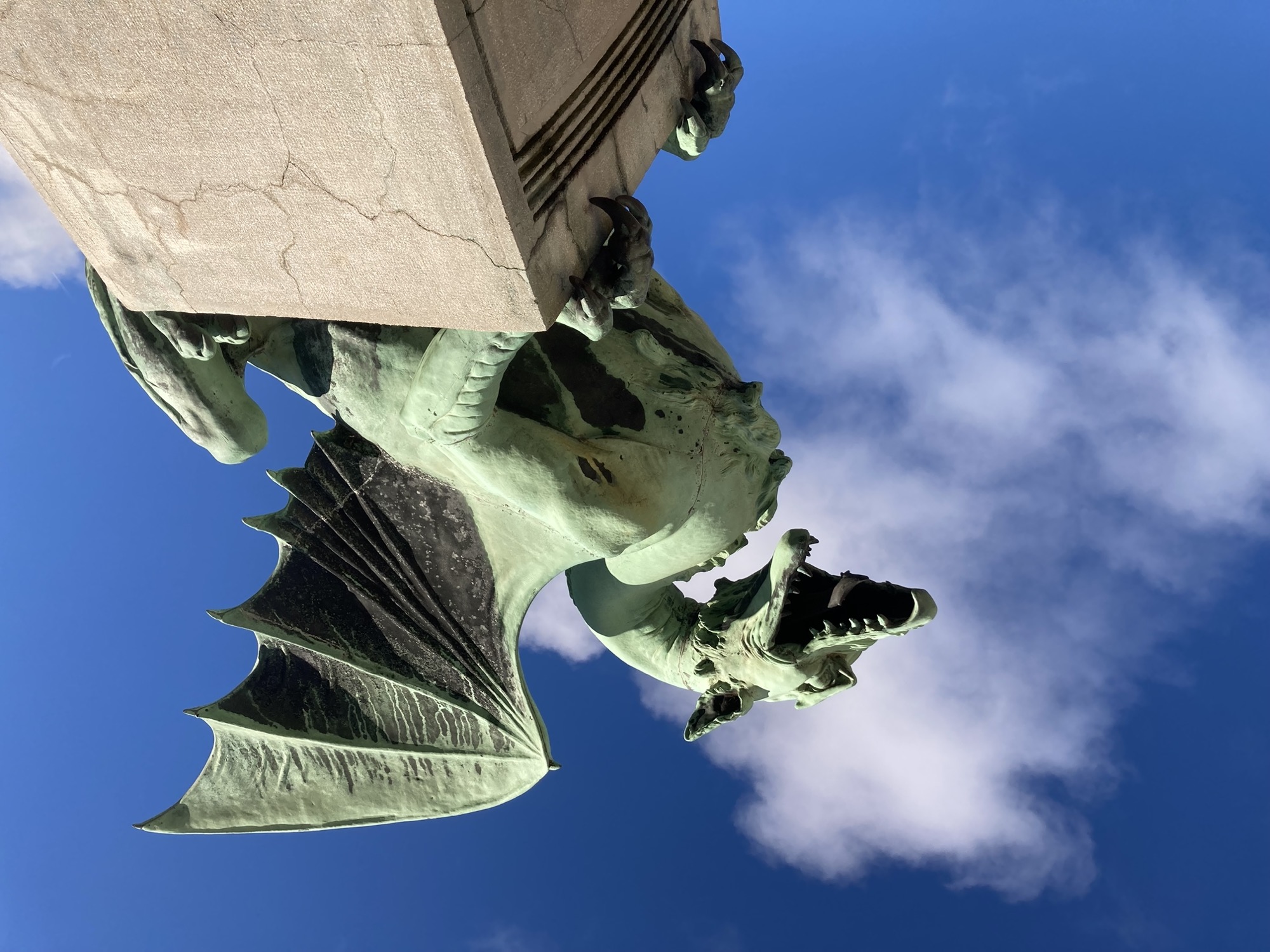 Statue eines Drachens mit aufgesperrtem Maul und gespreizten Flügeln, die aufrecht auf einem Sockel sitzt vor blauem Himmel mit einem luftigen Wölkchen