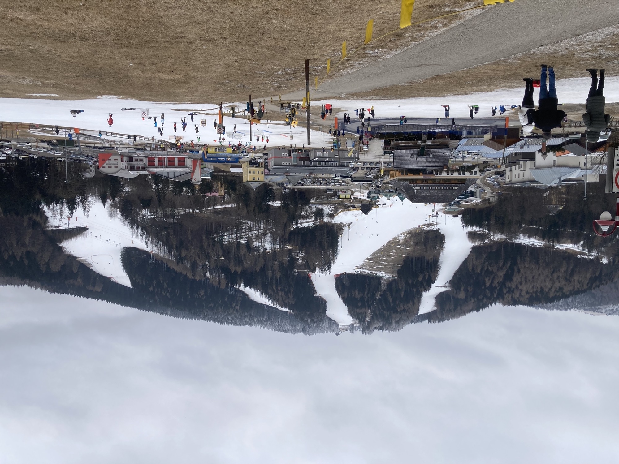 Ausblick auf einen Berg mit drei beschneiten Pisten, dazwischen Nadelbäume, nur bei den obersten sind die Spitzen auch weiß angehaucht, im Vordergrund eine flache Schneefläche für Kinder, ganz vorne ist der Boden braun und schneefrei