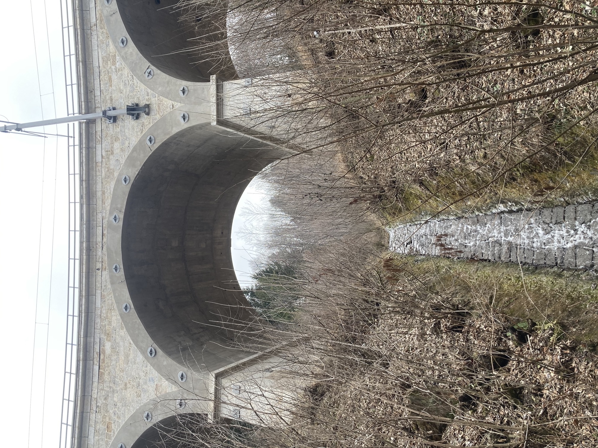 Ausblick von unten auf ein Eisenbahnviadukt, zwischen den massiven Säulen spannt sich ein Rundbogen, in der Mitte fließt ein kleines Bächlein steil bergab