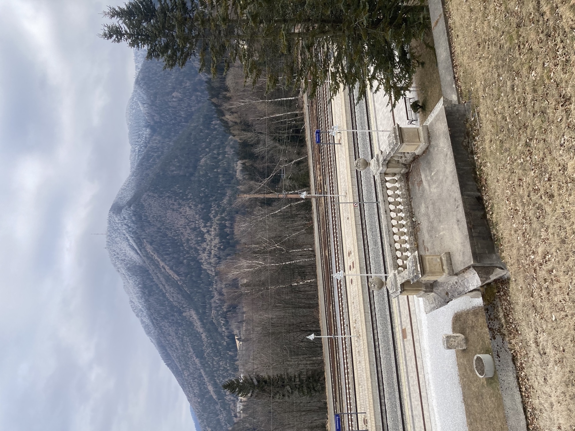 Bahnhof Semmering, vorne die Rückansicht des Ghega-Denkmals, mittig die Bahnsteige, im Hintergrund ein Berggipfel, der mit Schnee bezuckert ist (aber nur ganz oben)