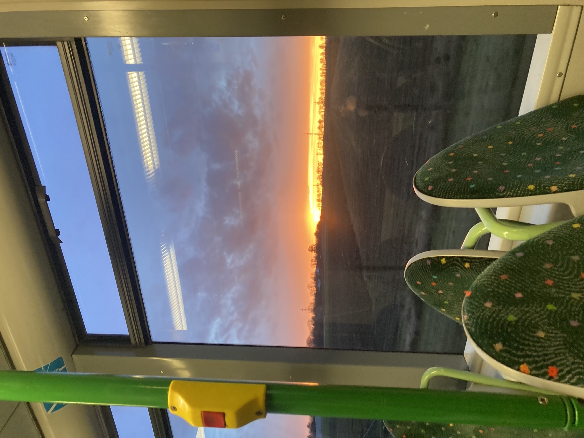 das Bild ist aus der fahrenden Straßenbahn aufgenommen, im Vordergrund eine Stange mit Halteknopf und grün gemusterte Sitze, durch das Fenster ist zu sehen, wie hinter einem Feld die Sonne untergeht, die Sonne beleuchtet nur noch einen schmalen Streifen des Himmels