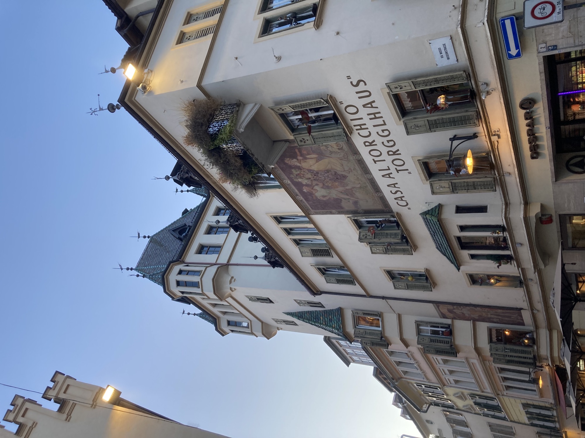 ein dekoratives Gebäude mit Erkern, Türmchen und grünen Fensterläden und Dachschindeln, ein Gemälde auf Höhe des dritten Stocks zeigt zwei Menschen, die eine überdimensionerte Weinrebe zwischen sich tragen, darunter der Schriftzug „Casa Al Torchio“ und darunter „Torgglhaus“