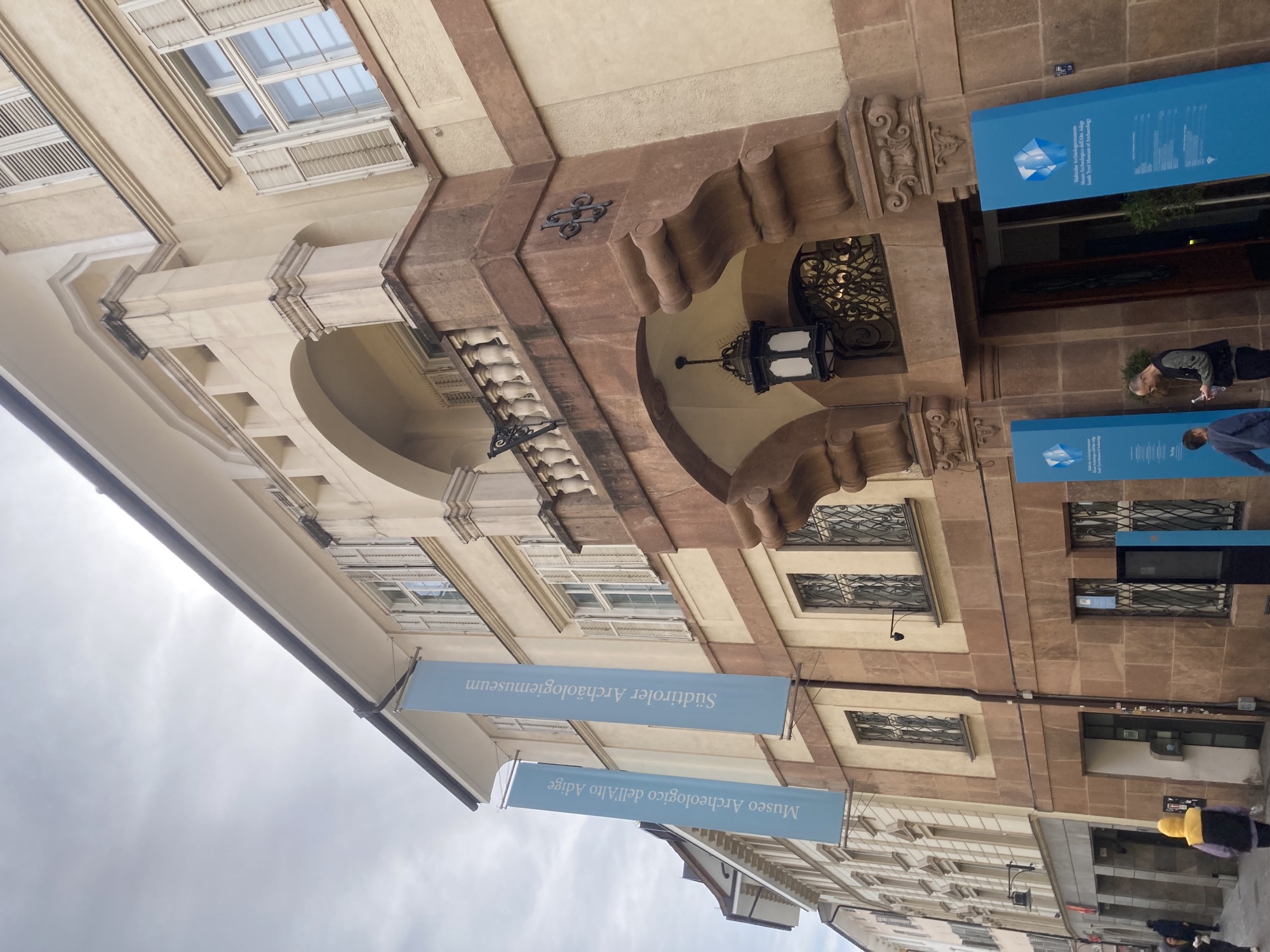 Eingang zum Südtiroler Archäologiemuseum, das Erdgeschoss ist mit großen braun gemusterten Marmorfliesen verkleidet, über dem Eingang hängt eine einzelne Lampe, darüber ein dekorativer Balkon mit einer Säulenbrüstung