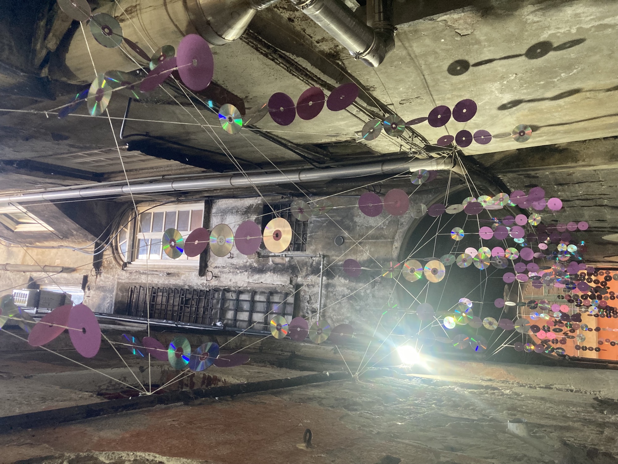 Kunstinstallation in einer sehr schmalen Gasse, zwischen heruntergekommenen Hauswänden sind Schnüre gespannt, an denen CDs aufgehängt sind, ein Scheinwerfer strahlt in die CDs hinein und sorgt somit für Lichteffekte
