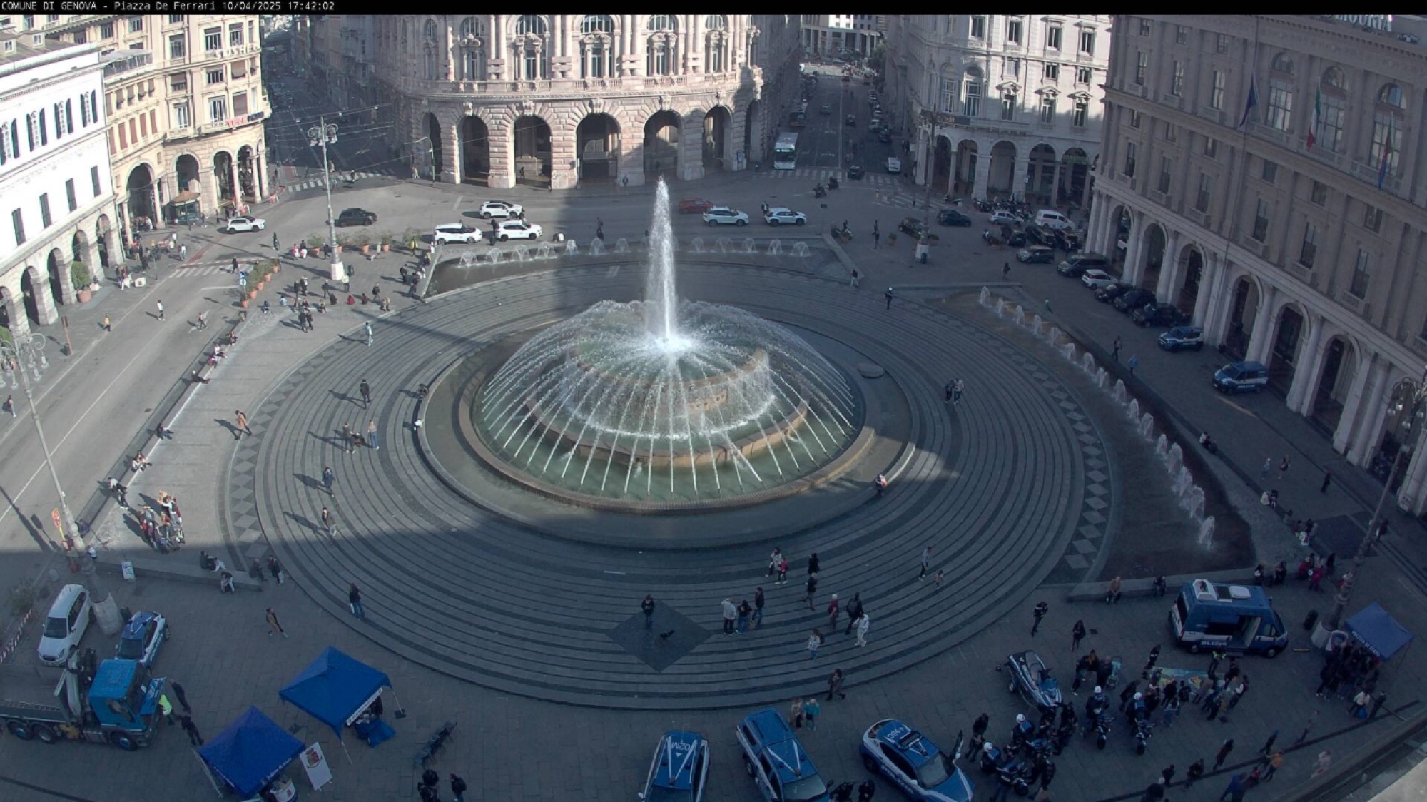 Webcam-Screenshot des großen Springbrunnens auf der Piazza De Ferrari, zentral im Bild ist der Springbrunnen gerade noch von Sonne beleuchtet, während der Großteil des Platzes schon im Schatten liegt, unten im Bild sind einige blau-weiße Polizeifahrzeuge zu sehen, zwischen diesen und dem Brunnen eine karoförmige Markierung auf dem Boden, in dieser steht eine Person mit einem Hund
