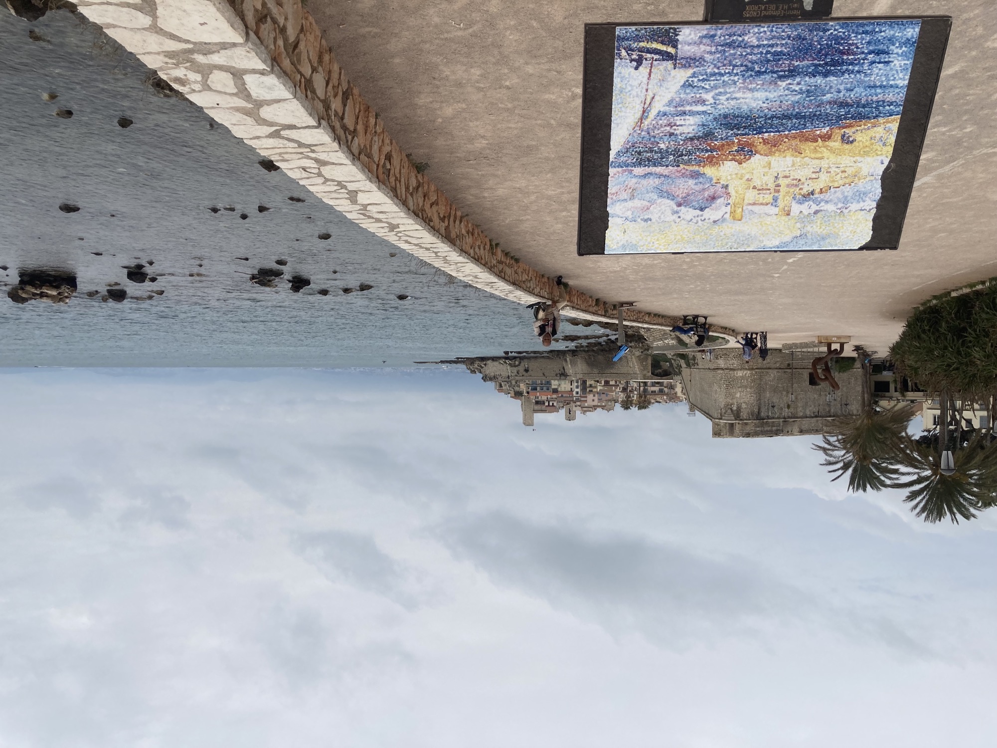 Promenade in Antibes, rechts das Meer, links vorne ein gemaltes Bild der Altstadt, die im Hintergrund in real zu sehen ist, das Bild ist deutlich farbiger als der echte graue Himmel, es zeigt die Altstadt in hellen gelb-braun-beige-Tönen und daneben das Meer in blau und violett