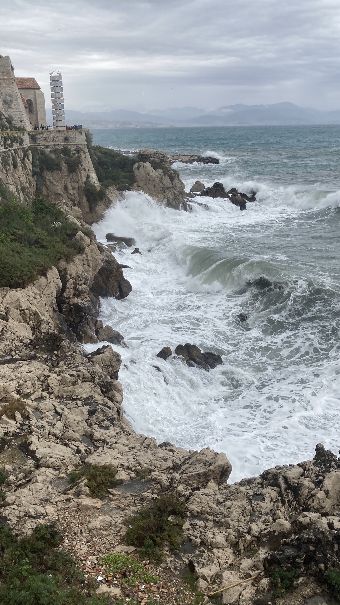 Wellen an der Steilküste unter der Promenade, das Meer rauscht sehr weiß, eine Welle bricht sich gerade im Anlauf auf die Klippen, die Berge im Hintergrund sind durch den Nebel nur zu erahnen