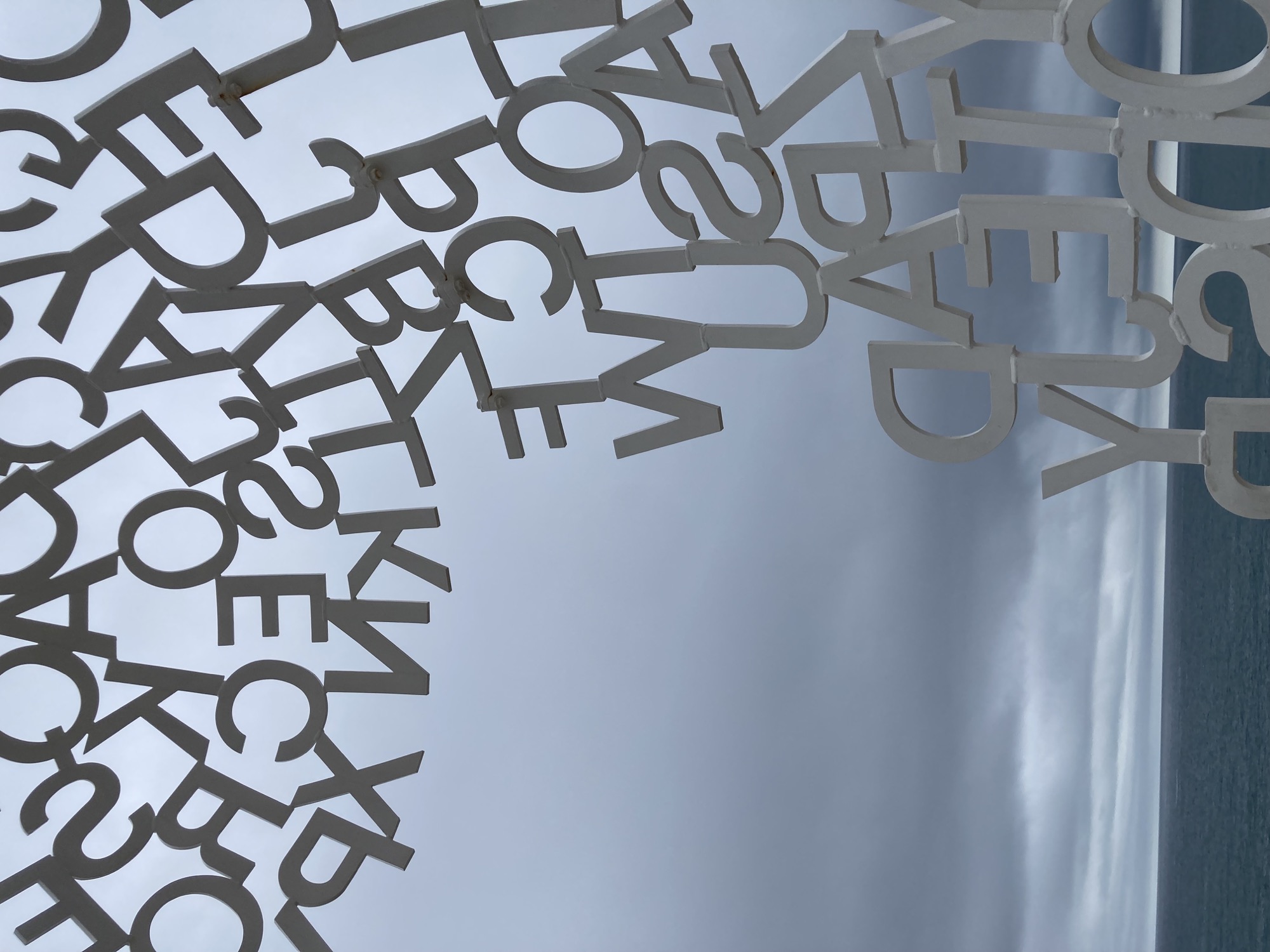 Detailaufnahme der Skulptur, es sind einzelne Buchstaben zu sehen, die zusammen die Struktur bilden, im Hintergrund grauer Himmel und dunkeklblaues Meer