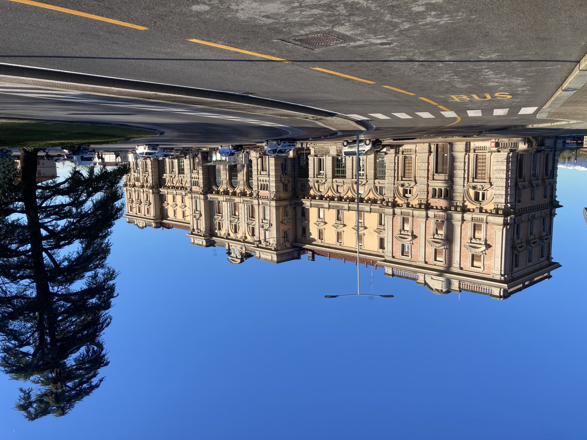 Bahnhofsgebäude von Cuneo, ein lang gezogenes Gebäude mit einem Mittelportal mit drei Toren und zwei Nebenflügeln an den Seiten, rechts im Bild ein großer Baum, vor dem Bahnhof stehen einige Autos