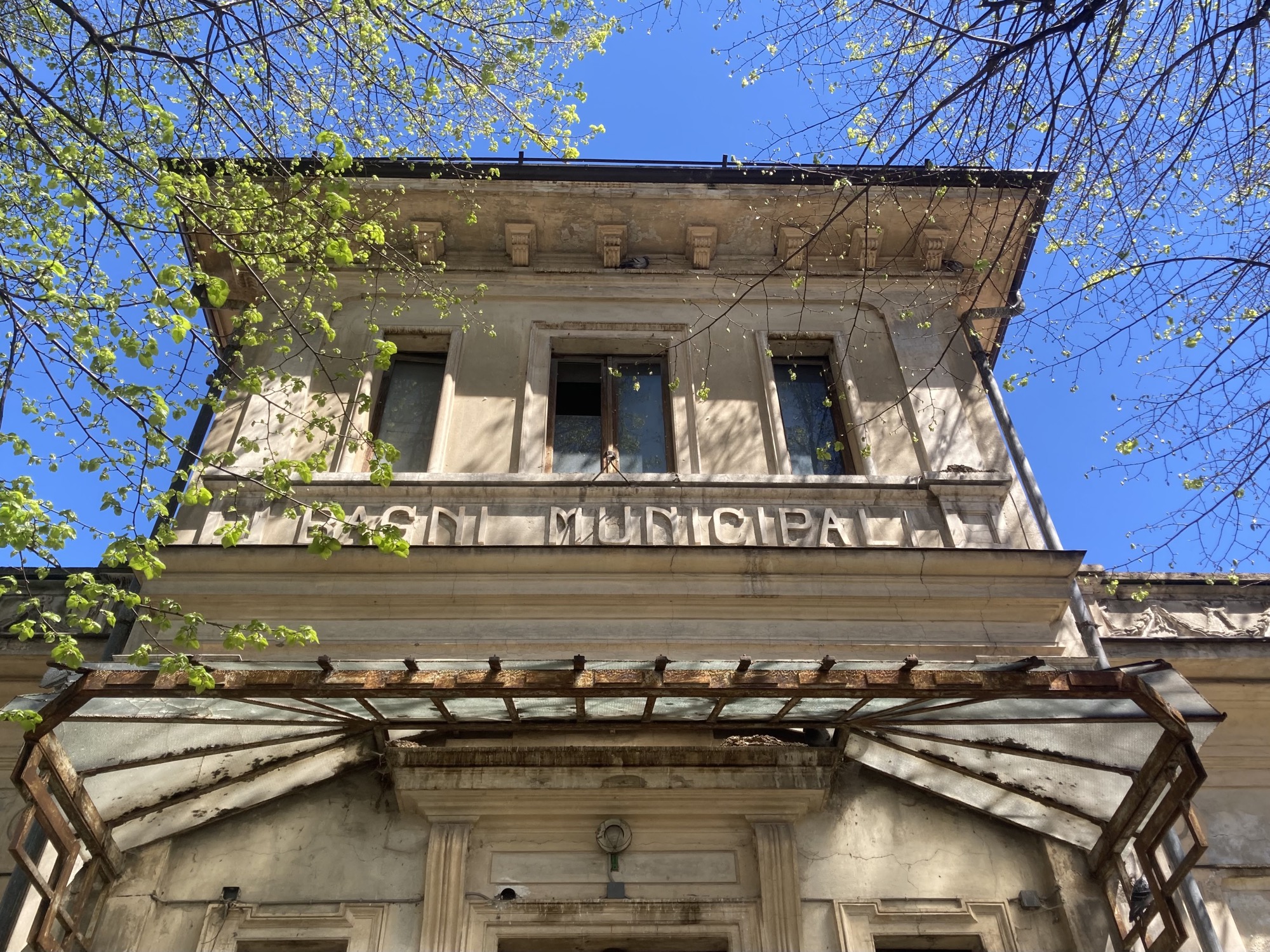 Blick nach oben auf die Fassade eines Hauses, über dem Eingang hängt ein schmutziges Glasdach, das wir ein Fächer angeordnet ist, darüber an der Wand der Schriftzug „Bagni Municipal“