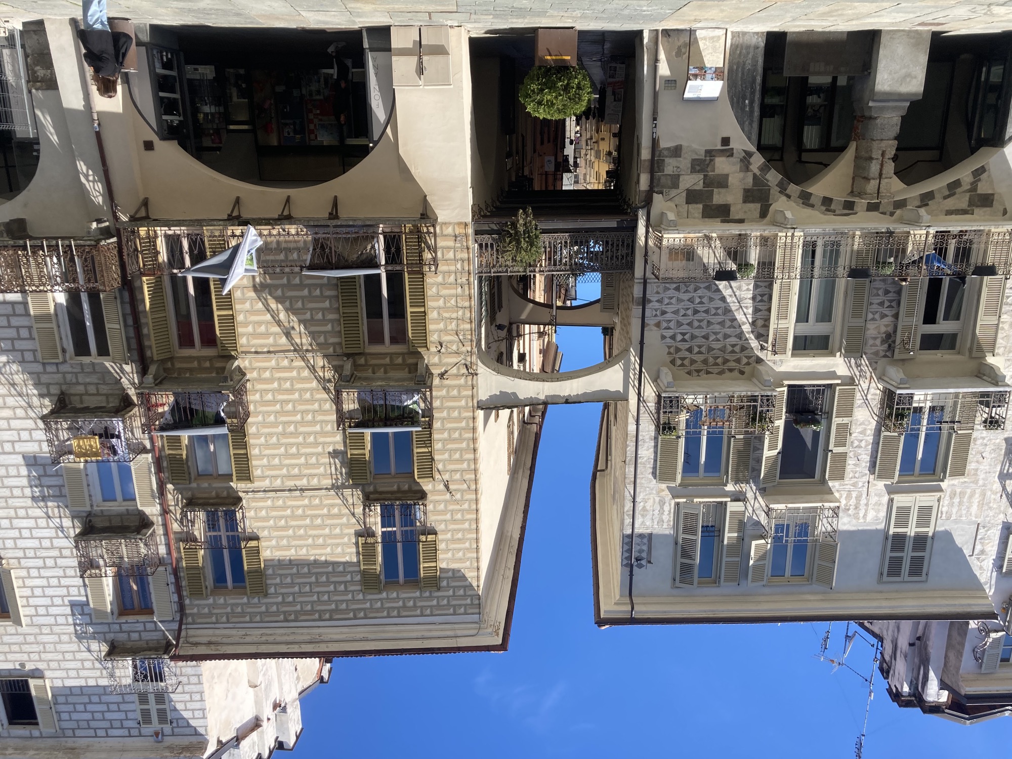 Häuserfassaden an der zentralen Straße von Cuneo, zwischen zwei Häuserfronten mit Balkonen und Fensterläden verschwindet eine kleine Gasse, die von Stützstreben überspannt wird