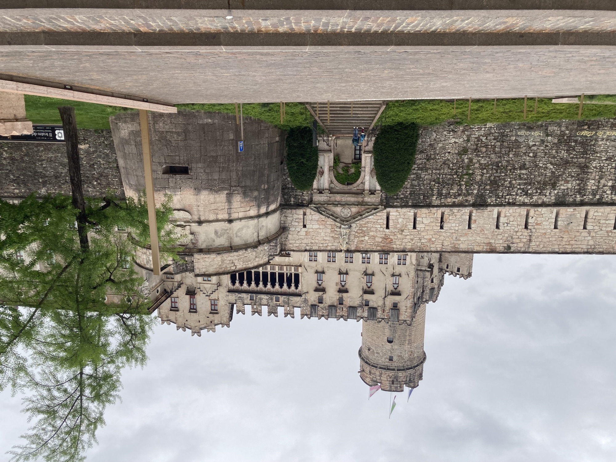 Eingang zu einer imposanten Burg, über dem Torbogen ein imposantes Gebäude mit einem Säulenbalkon, dahinter ein Turm, auf dem drei Flaggen wehen