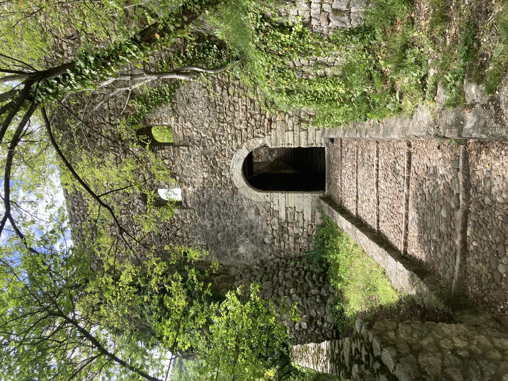 Weg zu einem Befestigungstor, Weg und Mauer sind aus Steinen gebaut, links und rechts überspannen Bäume und Sträucher den Weg und die Mauer