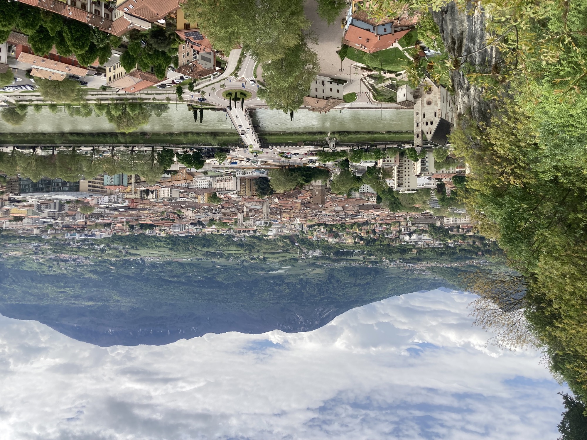 Ausblick auf den Fluss Adige/Etsch und dahinter die Stadt Trento, links eine Felsküste und darunter die Kirche vom oberen Bild, mittig ein Kreisverkehr, der auf die Brücke vom oberen Bild führt, ganz im Hintergrund Berge mit Nadelwald
