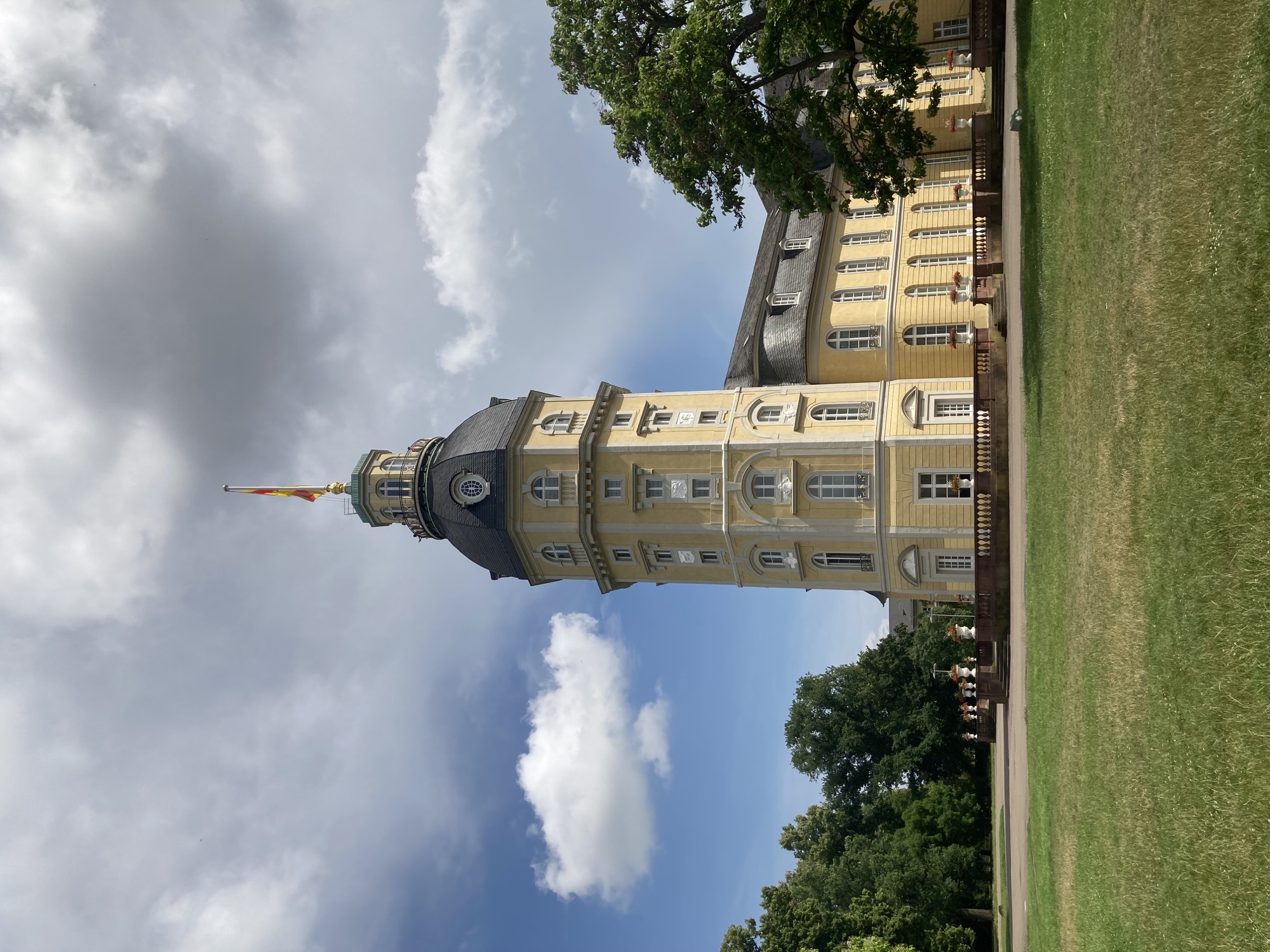 Turm im Schlossgarten, ein gelb gestrichenes Gebäude mit mehreren Stockwerken und weißen Verzierungen an den Fenstern, darauf eine graue Kuppel mit einer Flagge an der Spitze, dahinter bewölkter Himmel