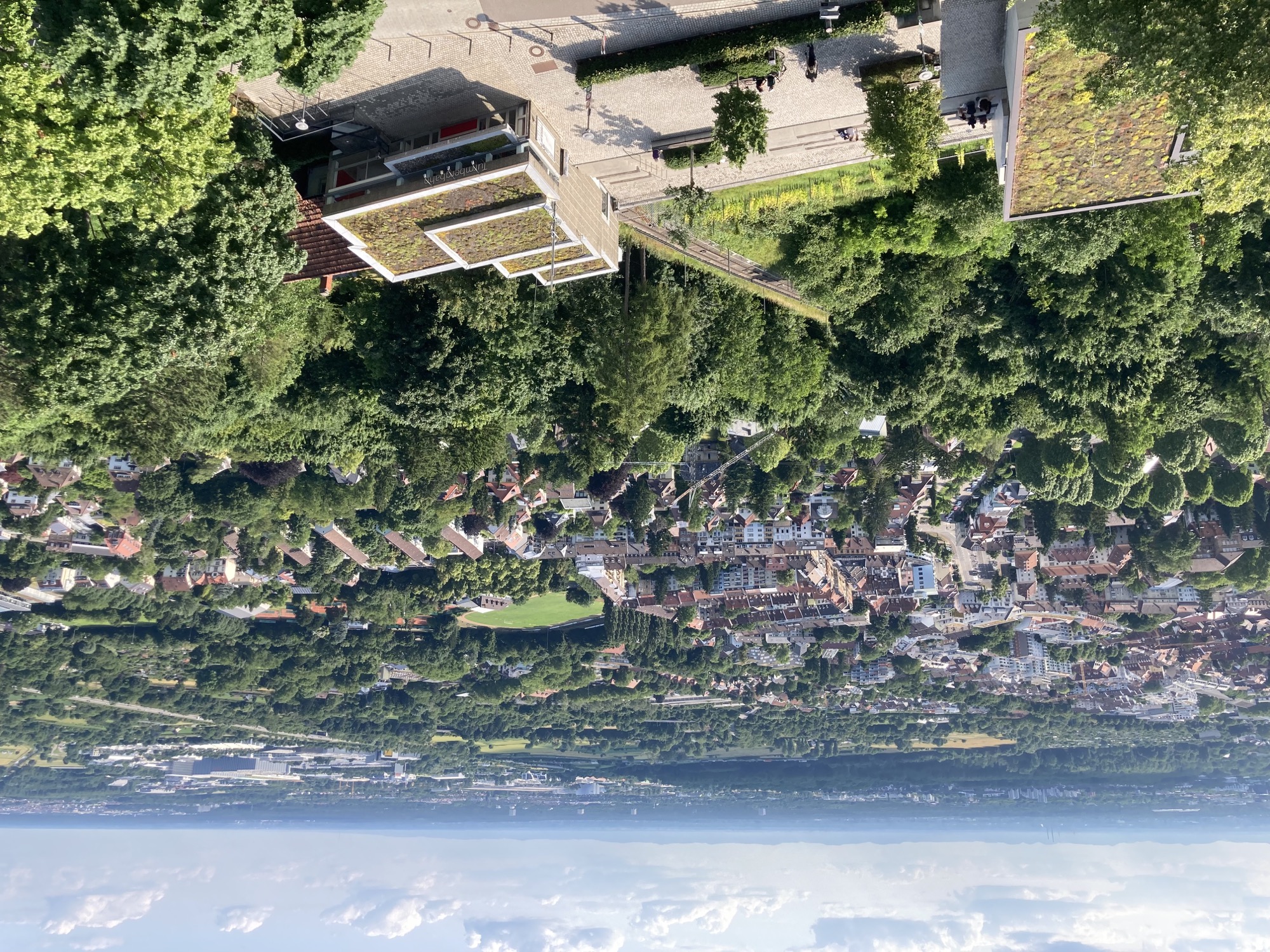 Ausblick von oben, im Vordergrund eine Terrasse neben der Bergstation der Turmbergbahn, dahinter die Häuser von Durlach, viele Wald- und Grünflächen, die Ebene streckt sich bis zum Horizont, auf dem kleine Wölkchen herumschweben
