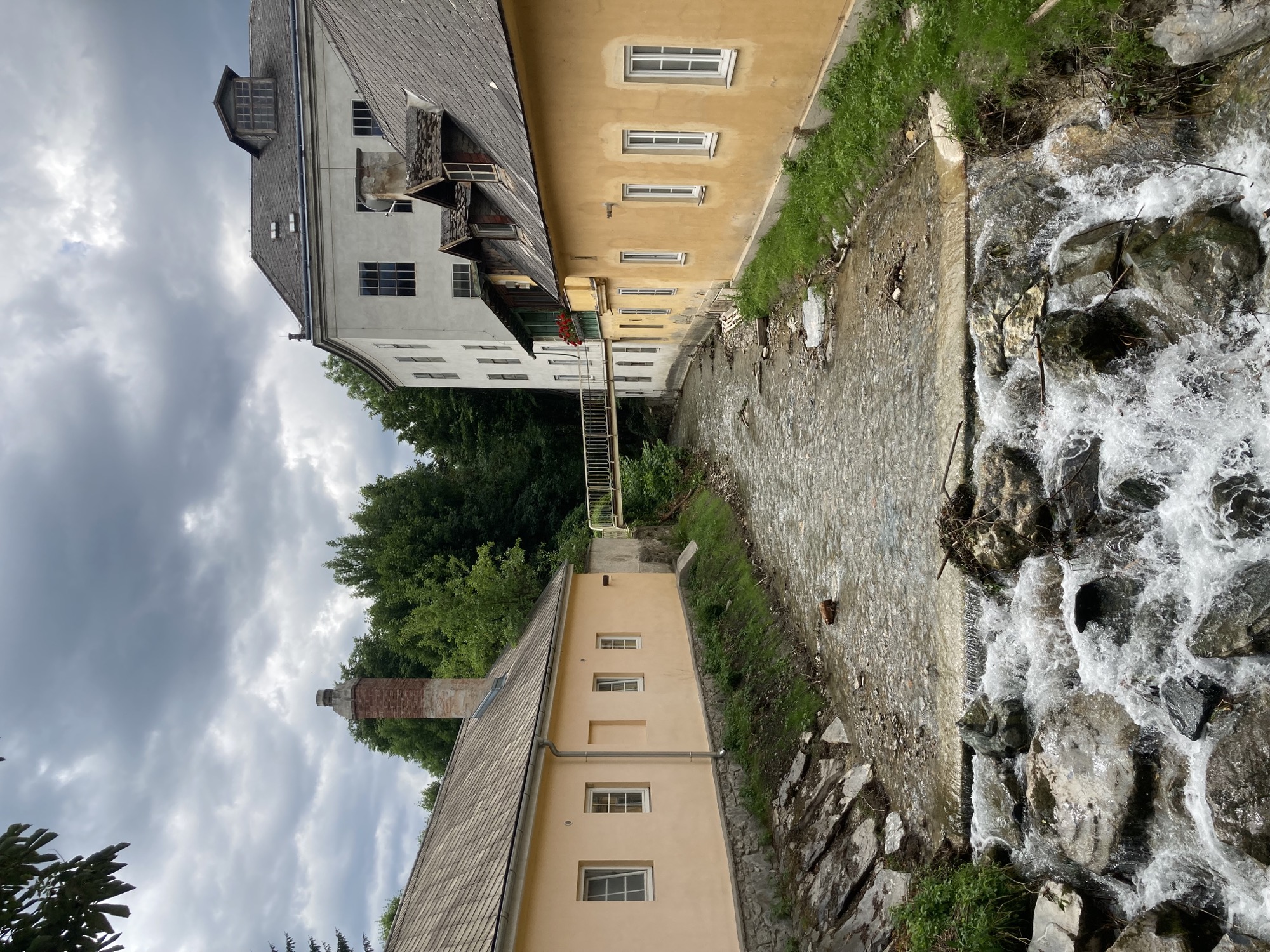 Blick auf einen Fluss, der zwischen zwei gelb gestrichenen Häusern verläuft, im Vordergrund des Bilds bricht sich der Fluss über einige Felsen bergab