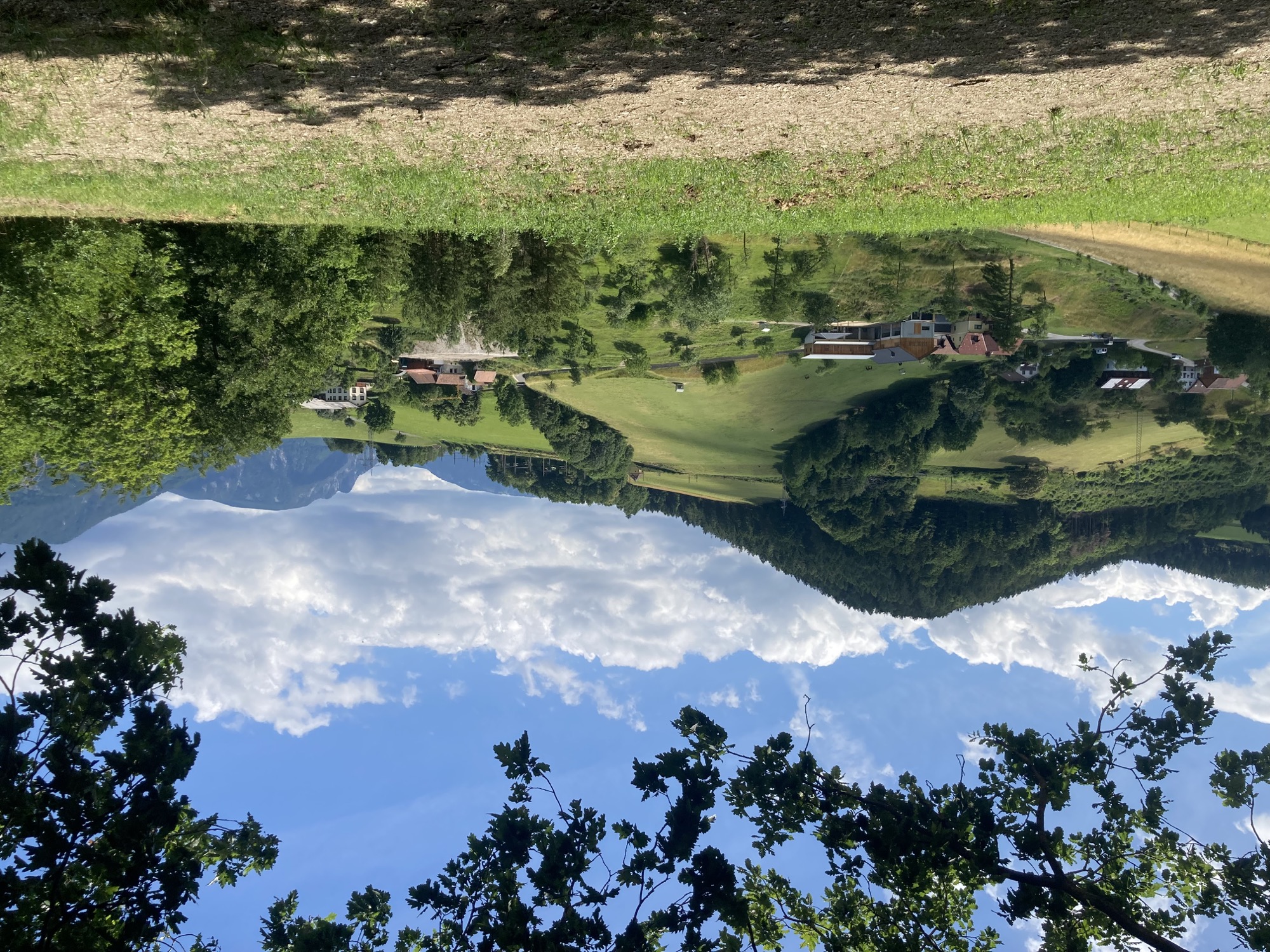 Foto einer idyllischen Berglandschaft, dunkelgrüne Wälder, hellgrüne Wiesen, dazwischen ein paar bäuerliche Gehöfte, höhere Berge und leicht bewölkter blauer Himmel im Hintergrund