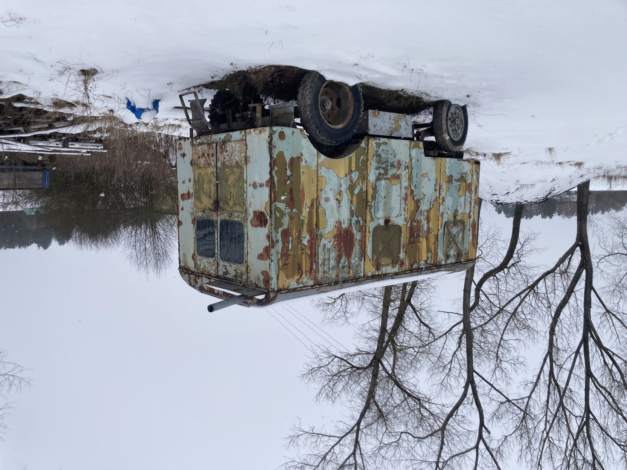ein Auto-Anhänger im Schnee, es wirkt etwas wie ein Zirkuswagen, verschiedene Farbschichten sind teilweise abgefallen, was dem Wagen eine gescheckte Farbe verleiht