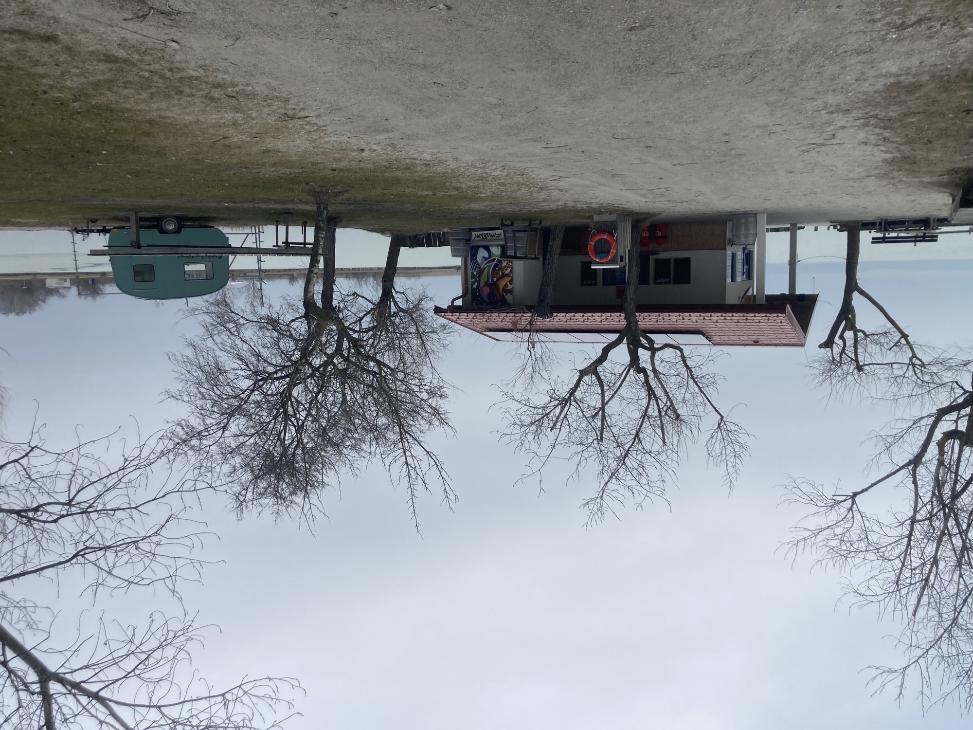 kleines, einstöckiges Haus am See, rundherum winterlich kahle Bäume, rechts steht ein kleiner Wohnanhänger, vor dem Haus hängt ein Rettungsring