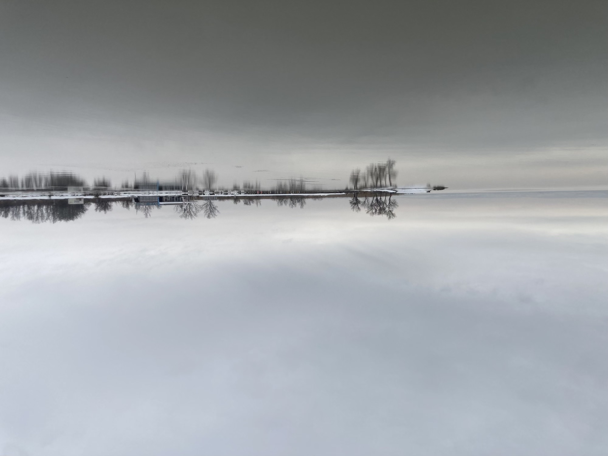 oben Himmel, unten See, beide in ähnlich grauer Färbung, weit hinten eine Landzunge mit Bäumen, die ins Wasser ragt, das Ufer ist von Schnee bedeckt