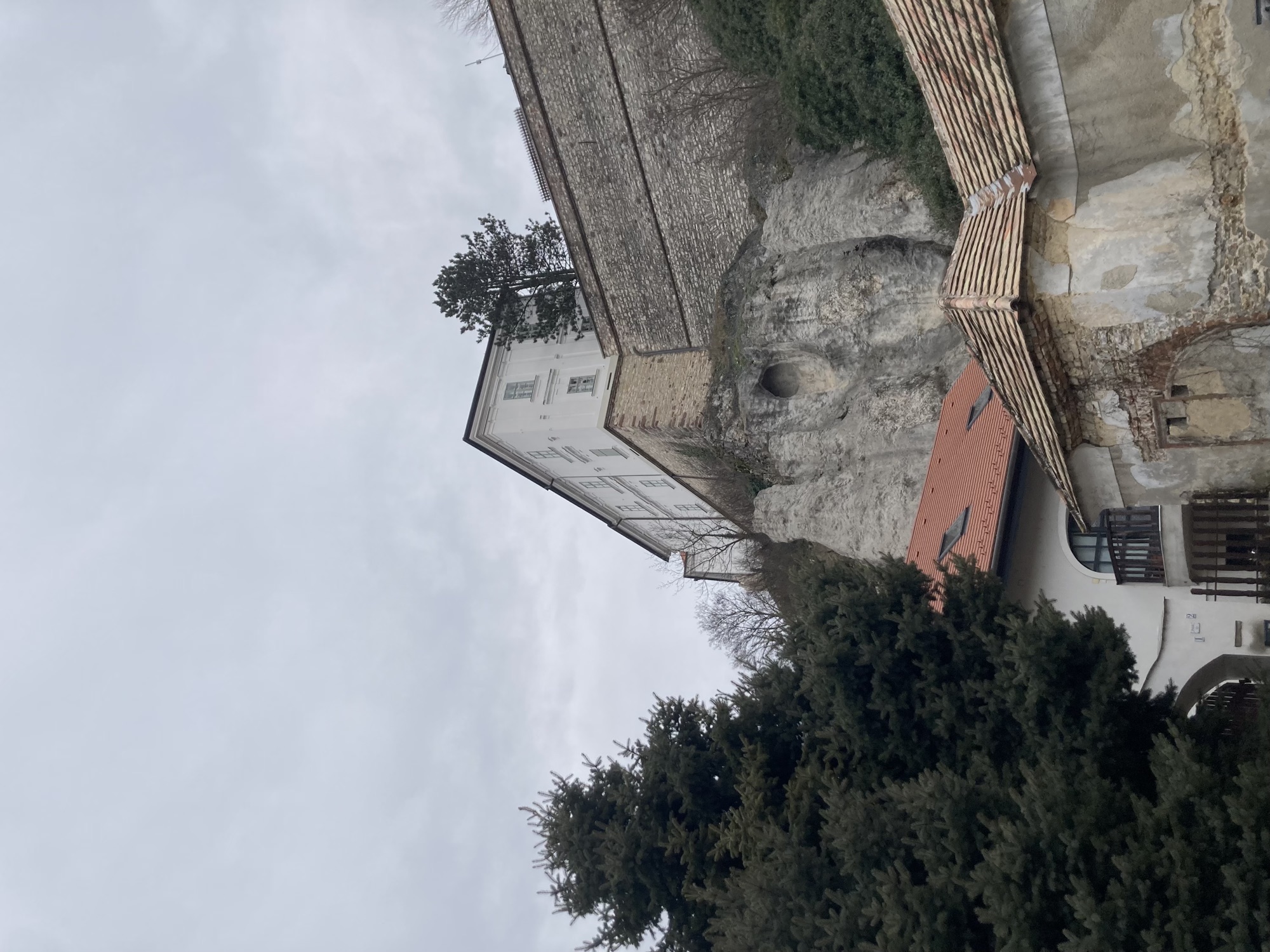 Blick nach oben auf den Burgberg, über einer Felswand und einer Ziegelmauer thront ein weißes, zweistöckiges Gebäude vor grauem Himmel