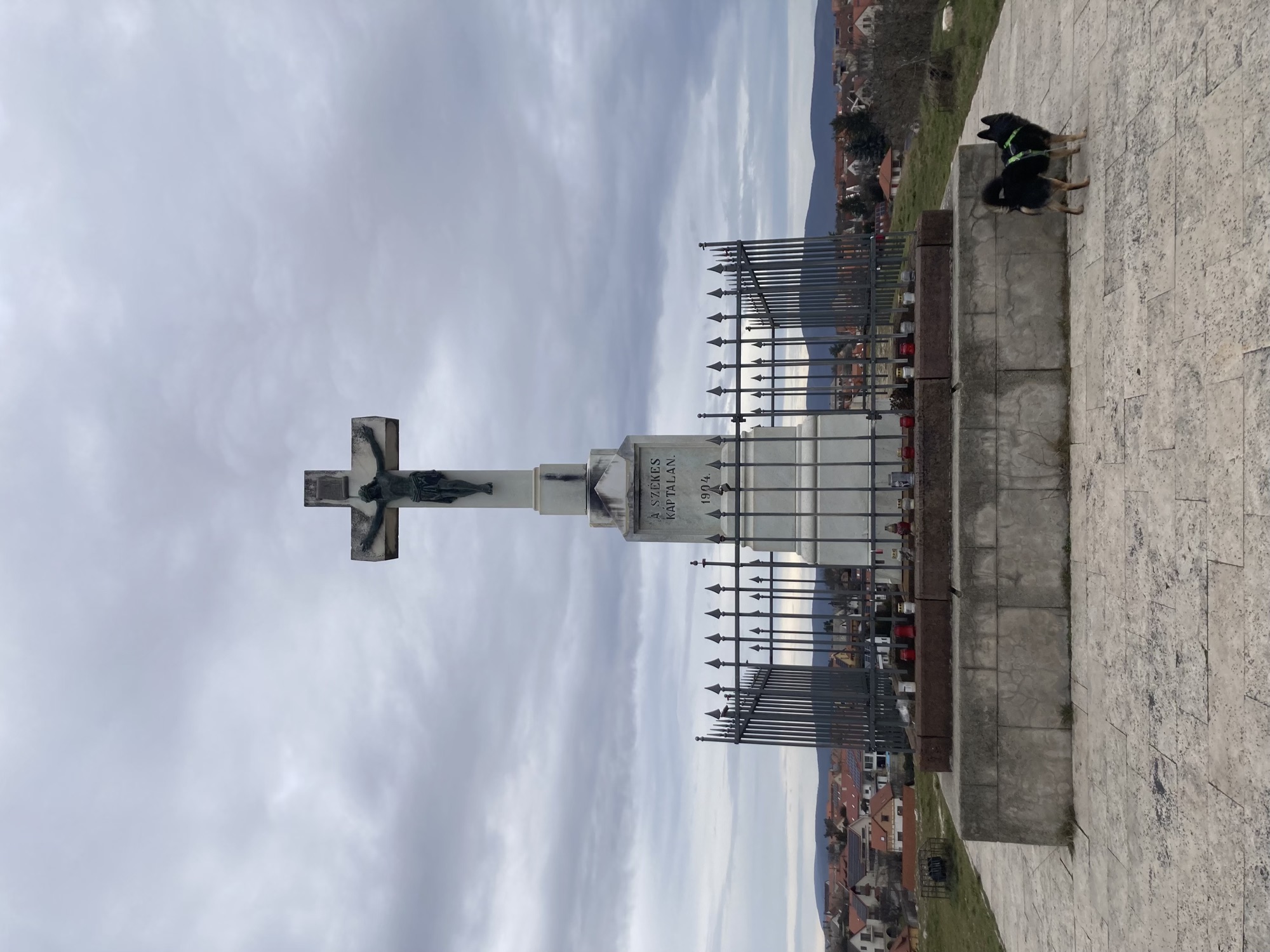 Jesus am Kreuz auf einem weißen Sockel, umringt von einem Zaun mit Spitzen oben, vor dem Zaun stehen einige Kerzen, rechts an der Ecke des Sockels schaut ein kleiner schwarzer Hund von der Kamera weg um die Ecke des Sockels