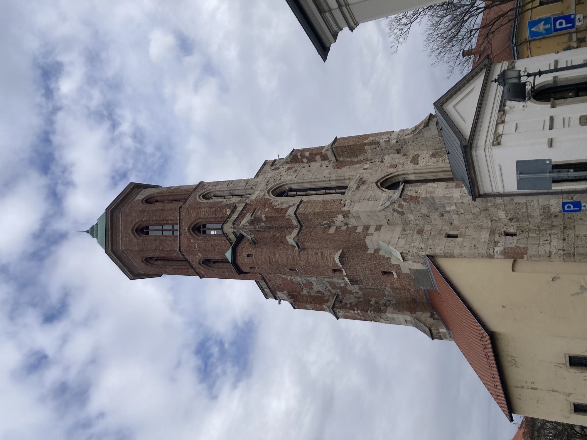 Kirchturm mit mehreren Stockwerken, die Wand bestand ursprünglich aus rötlichen Ziegeln, wurde aber an mehreren Stellen mit helleren Steinen ausgebessert, unten rechts führt ein weißes Portal mit einem grauen Spitzdach ins Innere