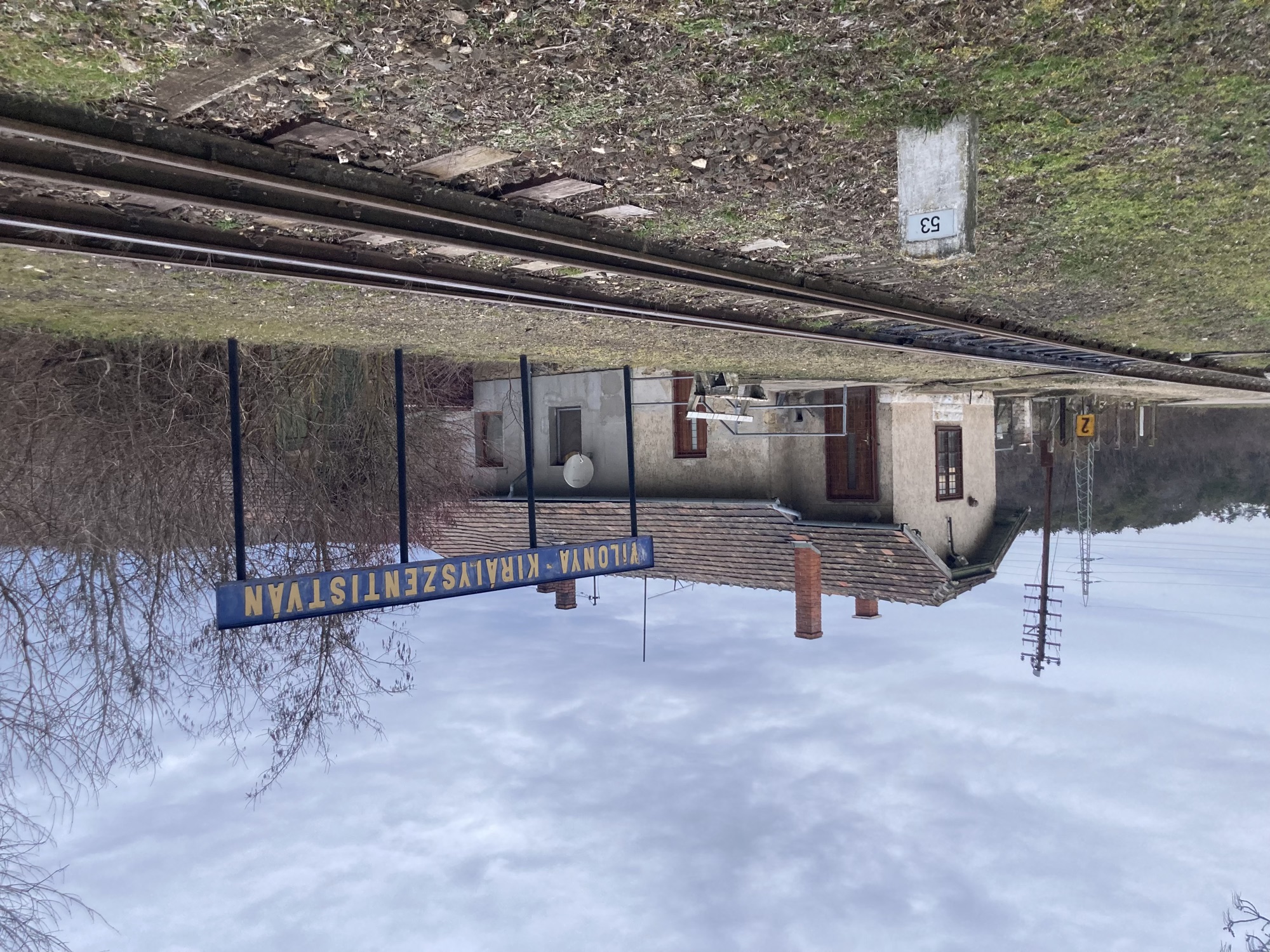verlassene Bahnstation, im Vordergrund ein Stein mit der Zahl 53 neben einem Gleis, dahinter ein breites blaues Stationsschild mit der gelben Aufschrift „VILONYA KIRÁLYSZENTISTVÁN“, dahinter das Stationshaus mit vier Rauchfängen auf einem altmodischen Ziegeldach