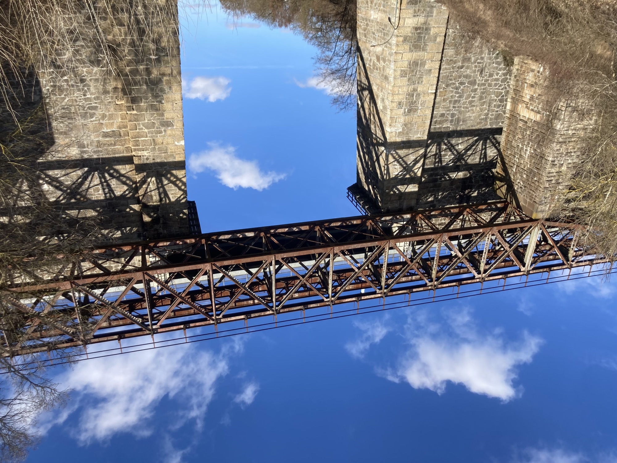 eine Brücke aus altem Metall verbindet zwei hohe Mauern aus alten Ziegeln, die Brücke ist deutlich rostig, ihr Muster zeichnet sich als Schatten auch auf den Mauern ab, dahinter blauer Himmel mit vereinzelten Wolken