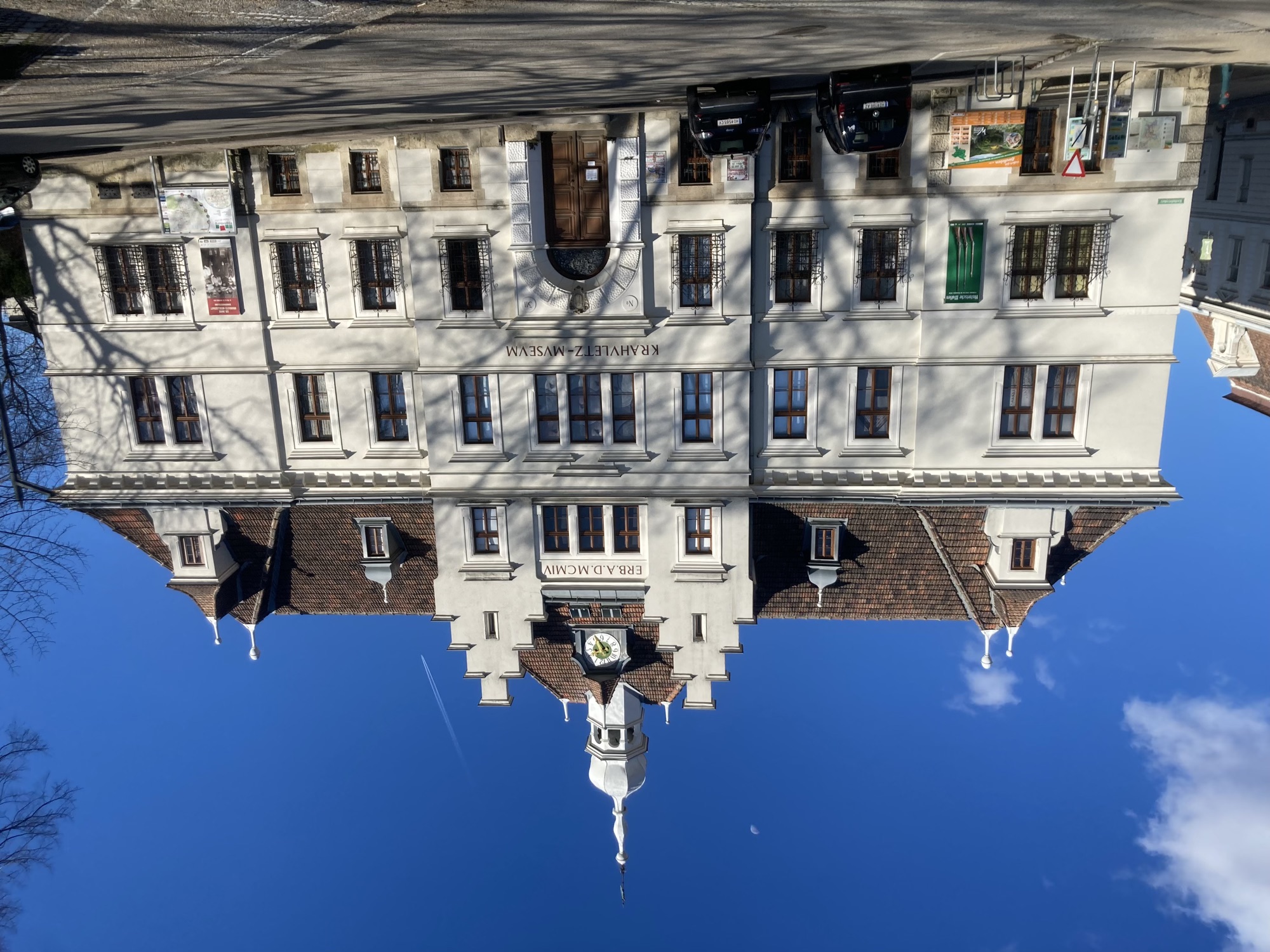 das Krahuletz-Museum, ein dreistöckiges, auf einer abfallenden Straße errichtetes Gebäude, im Erdgeschoss sind die Fenster also links höher und werden nach rechts immer niedriger, oben in der Mitte eine dekorative Uhr zwischen zwei Ziergiebeln, dahinter ein barockes Zwiebeltürmchen, dahinter blauer Himmel, links vom Turm ein halber Mond, rechts vom Turm ein Chemtrail Richtung Boden