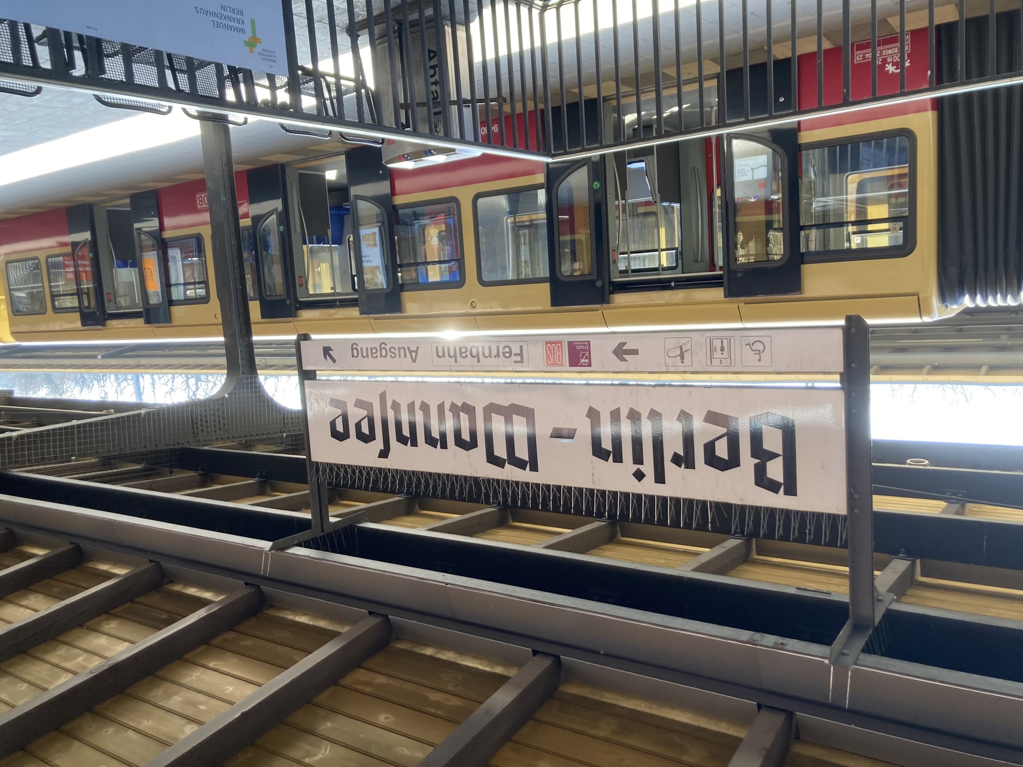 Bahnsteig der S-Bahn mit dem Haltestellenschild „Berlin-Wannsee“ in ebrochener Schrift, dahinter eine S-Bahn mit geöffneten Türen