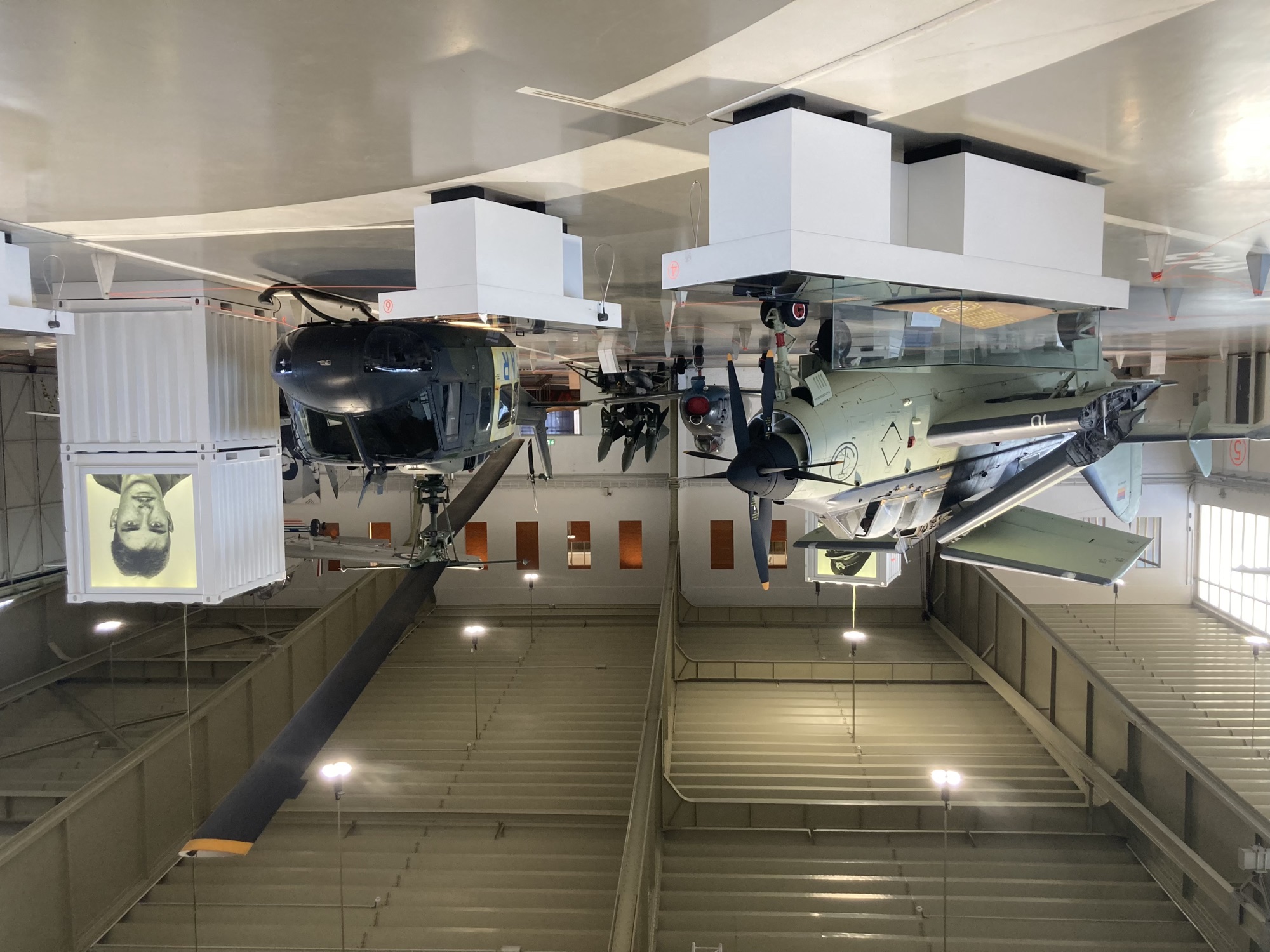 Ausstellung im Hangar, links ein hellgrünes Flugzeug mit zusammengeklappter Tragfläche und einem schwarzen Propeller, daneben ein schwarzer Helikopter