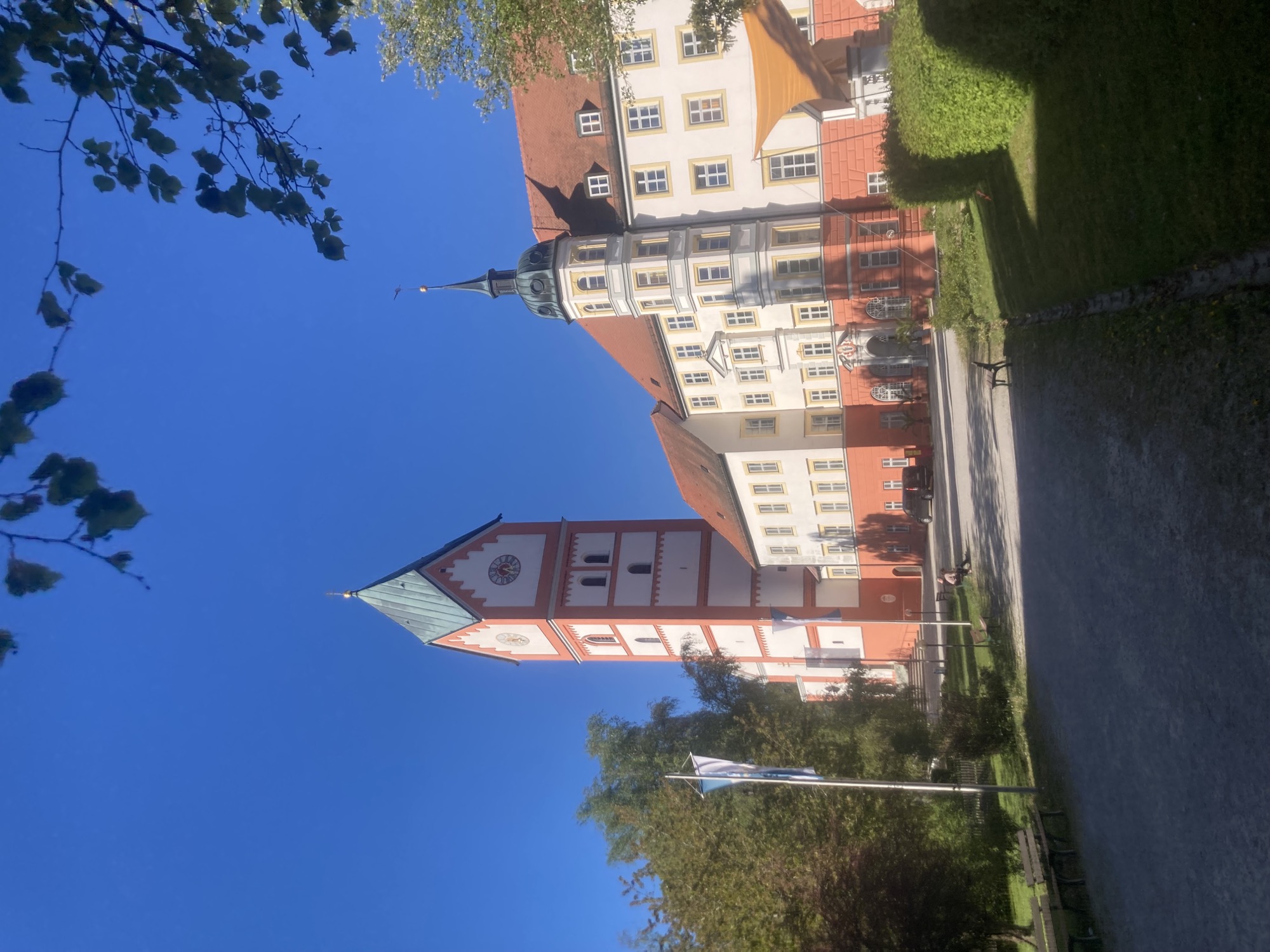 dekoratives Klostergebäude in weiß und hellrot inmitten eines Parks, links Kirchturm mit Turmuhr, rechts davon ein Wohngebäude mit einem Ecktürmchen