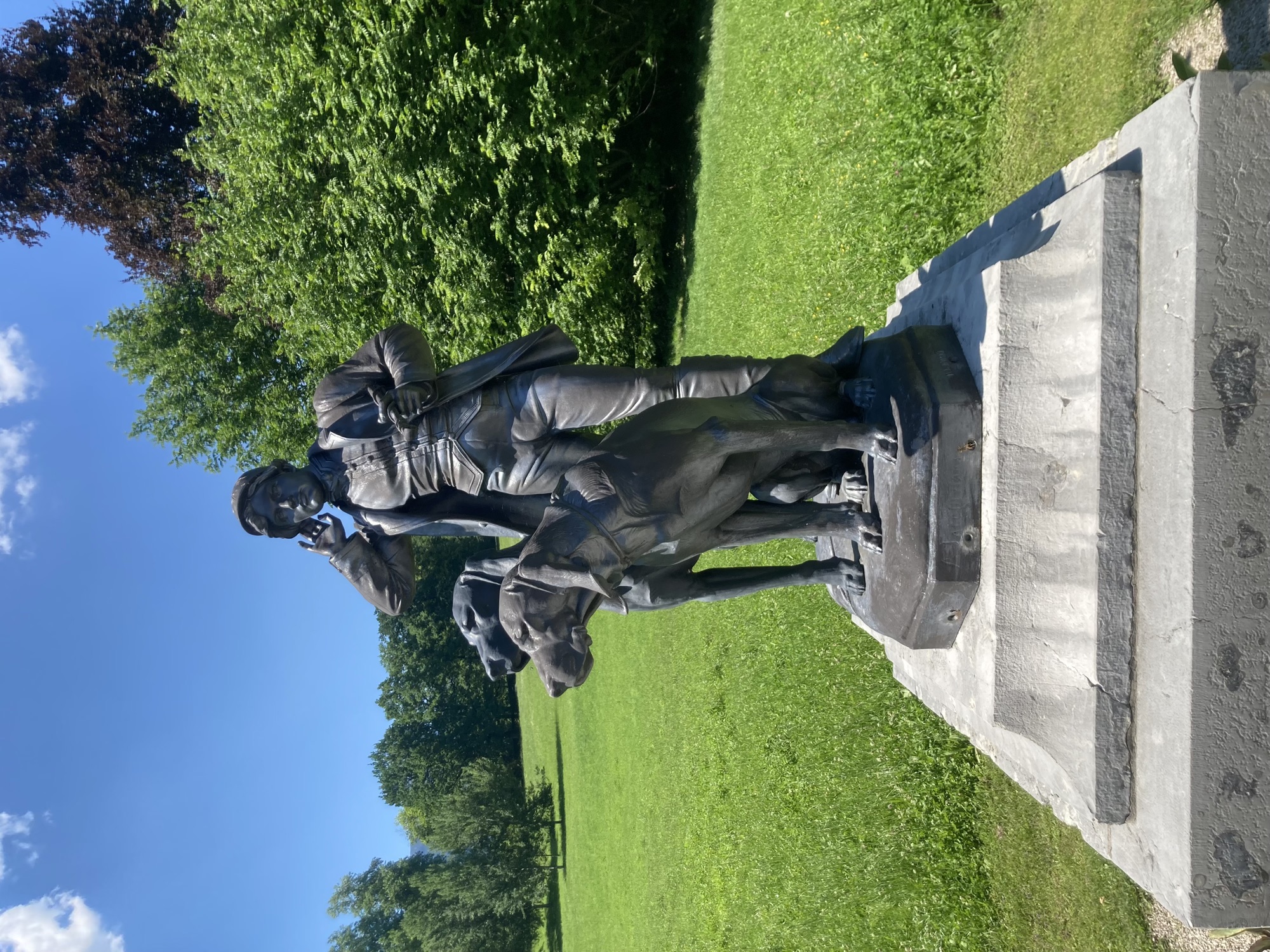 Metallstatue auf einem Steinsockel, ein Jäger hält die Hand ans Ohr, als würde er lauschen, neben ihm schauen zwei große Jagdhunde in dieselbe Richtung